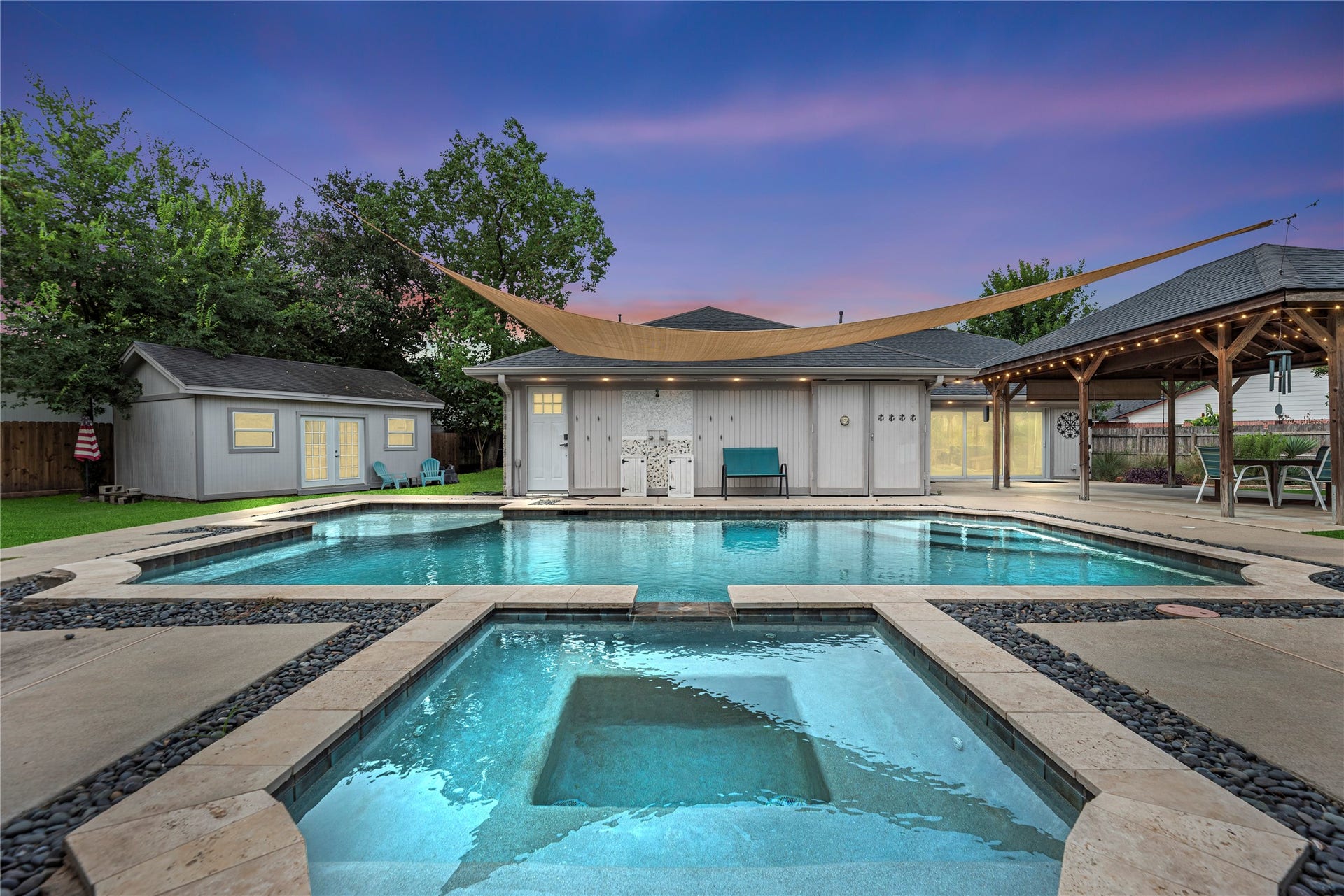 2418 Pebbledowne Circle, Sugar Land, TX 77478 Resort-style backyard showcase featuring an in-ground swimming pool with built-in spa, dark river rock surround, expansive concrete decking, large freestanding wooden gazebo, detached storage shed/workshop, and overhead sun shade sail.