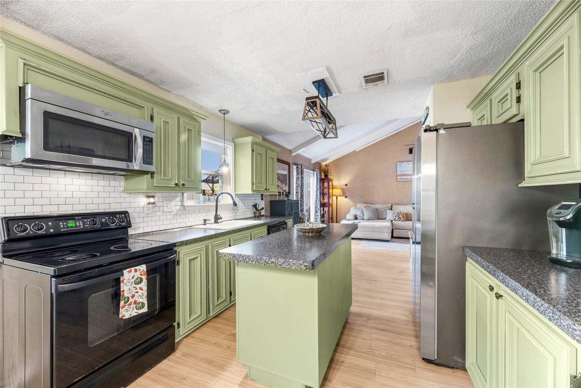Kitchen with green cabinetry, a center island, and stainless-finish appliances, with the living area visible beyond.