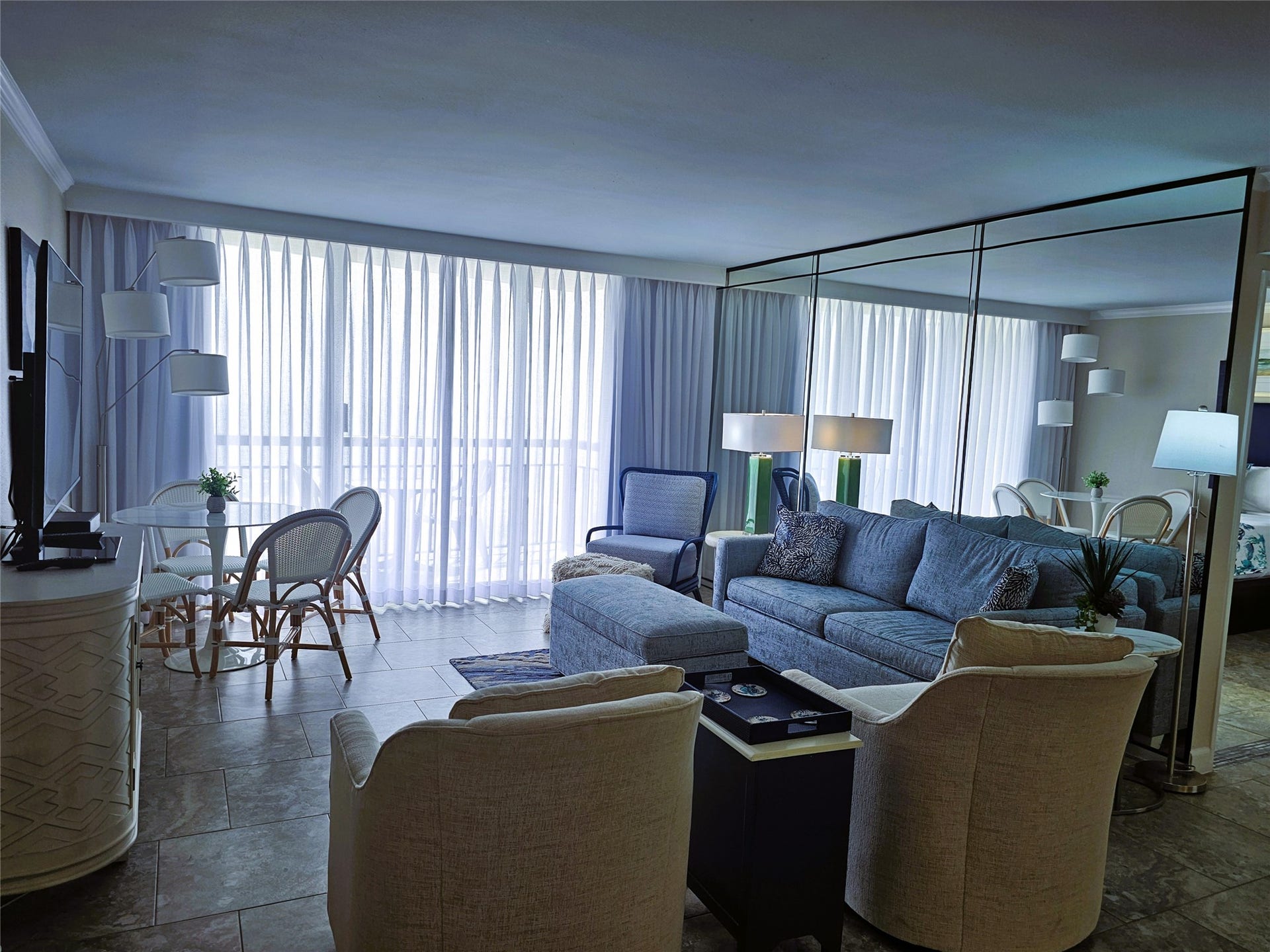 Bright and inviting living area with floor-to-ceiling windows, cozy seating, and a stylish mirrored wall enhancing the sense of space.