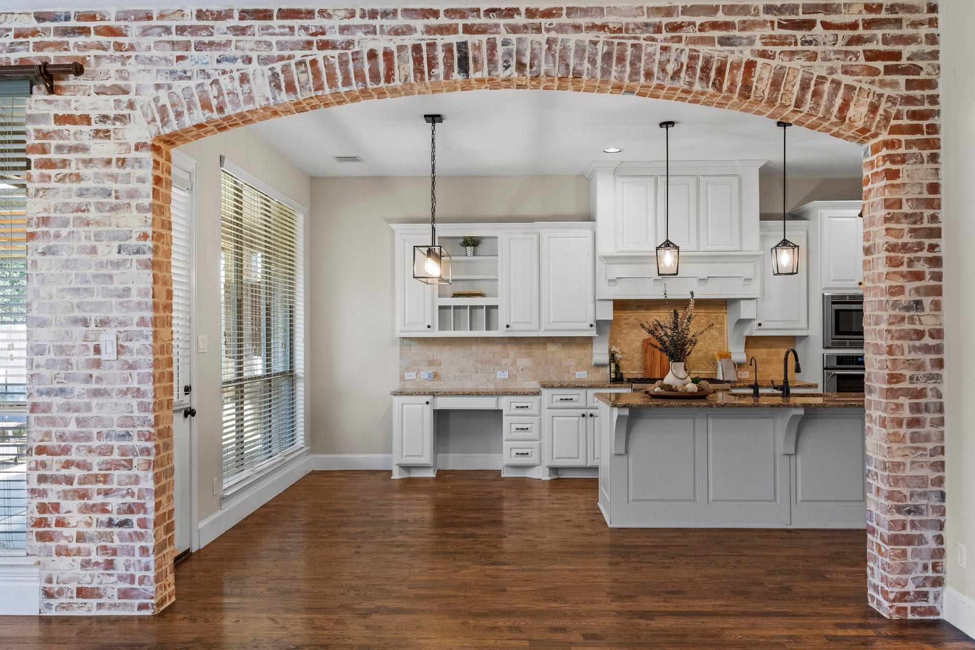 9238 Balsam Gap, Missouri City, TX 77459 A beautiful brick archway frames the spacious kitchen, featuring white cabinetry, granite countertops, a large center island with seating, pendant lighting, and rich hardwood floors. A charming architectural detail that creates a seamless connection between the kitchen and family room.