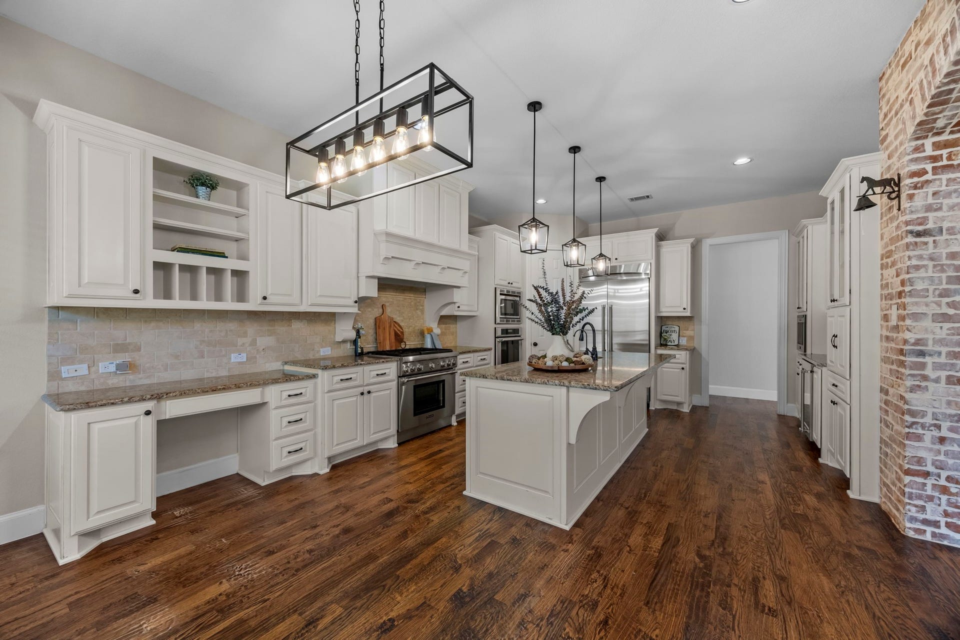 9238 Balsam Gap, Missouri City, TX 77459 chef's kitchen featuring beautiful white cabinetry, granite countertops, stainless steel appliances, and a large center island with sink. Custom vent hood, built-in desk area, decorative pendant lighting, rich hardwood floors, and charming brick accents add character and functionality to this inviting space.