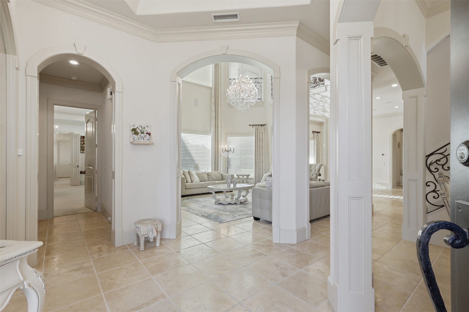 An open foyer highlights soaring tray ceilings, wide-cased arched passages, substantial baseboard moulding, and travertine tile flooring flowing seamlessly into the main living areas.