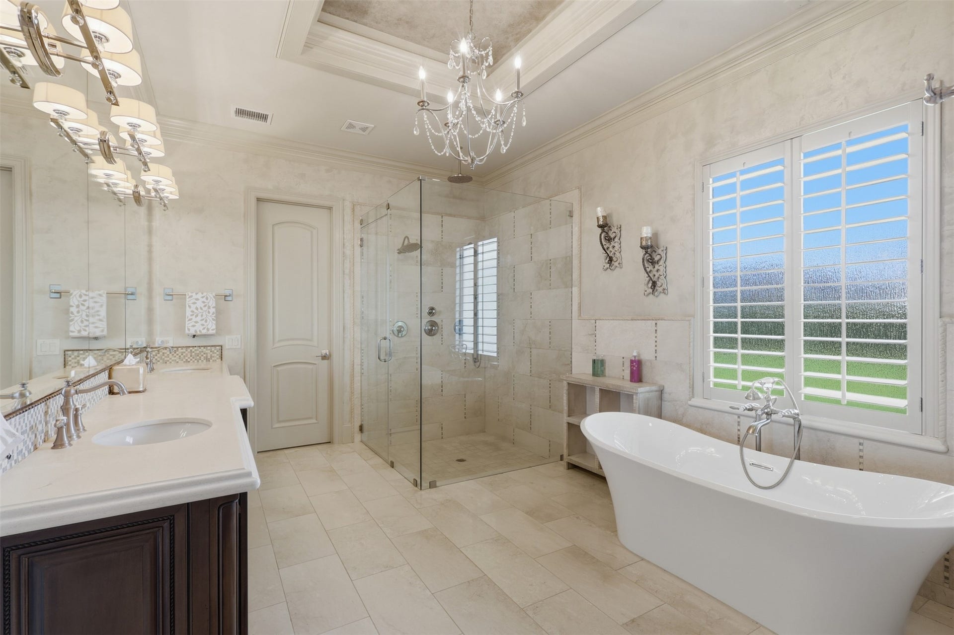 The home's primary bathroom features a crystal chandelier, dark-finish double vanity with stone countertops, travertine flooring, and a deep soaking tub.