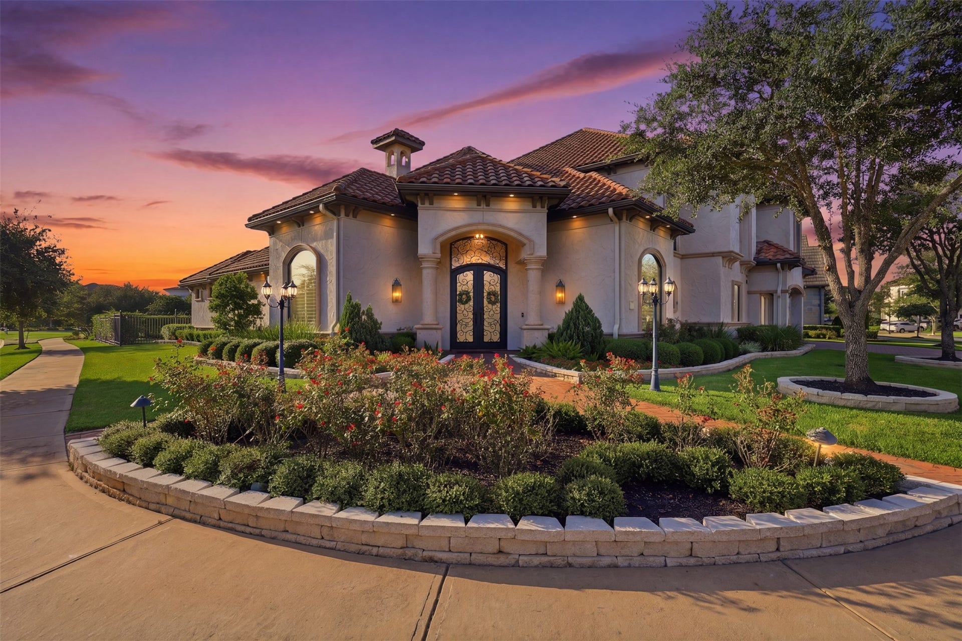 Symmetrical architectural lines are accented by ambient exterior sconce lighting, highlight corbels, decorative stone trim, and clay tile rooflines set against a twilight sky.