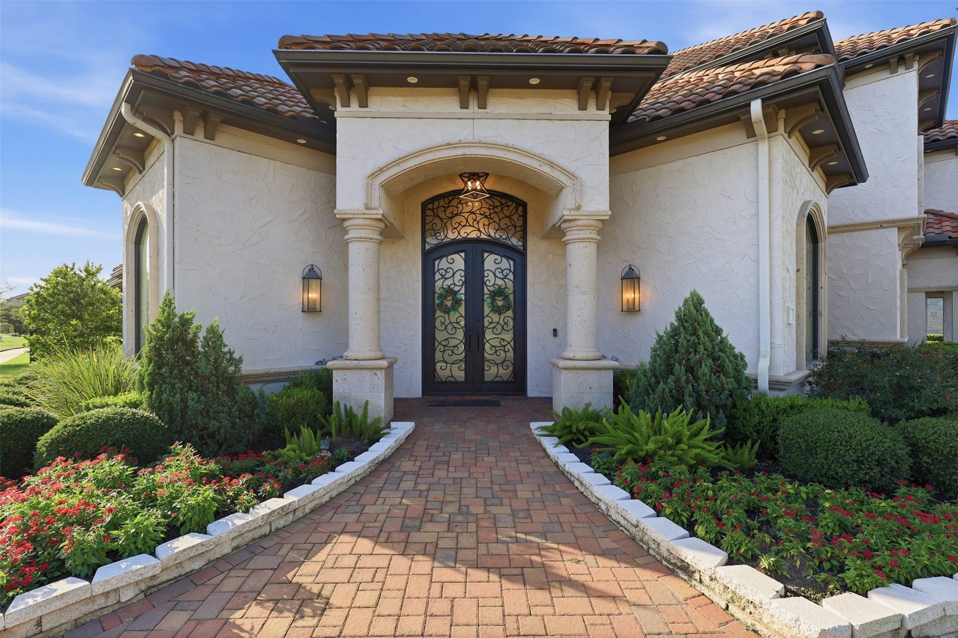 A grand portico framed by stone columns leads to a custom wrought-iron double door with ornate glass scrollwork, set against a classic Mediterranean tile roof and paver walkway.