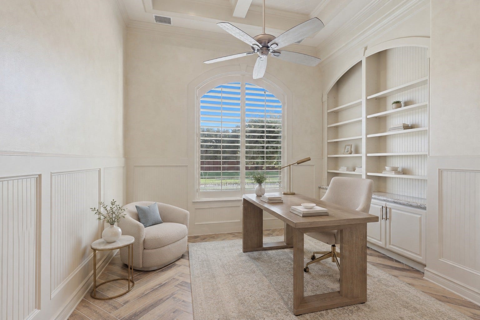 The study features custom floor-to-ceiling arched shelving, classic wainscoting trim, herringbone patterned wood tile flooring, and an arched window fitted with plantation shutters.**Virtually staged