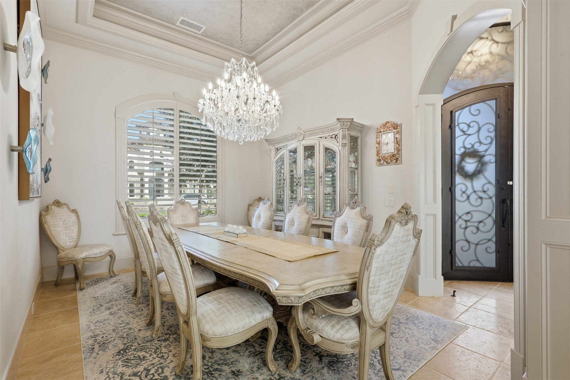 The formal dining space features a multi-tiered recessed tray ceiling, detailed crown moulding, a brilliant crystal chandelier, and an arched window with plantation shutters.
