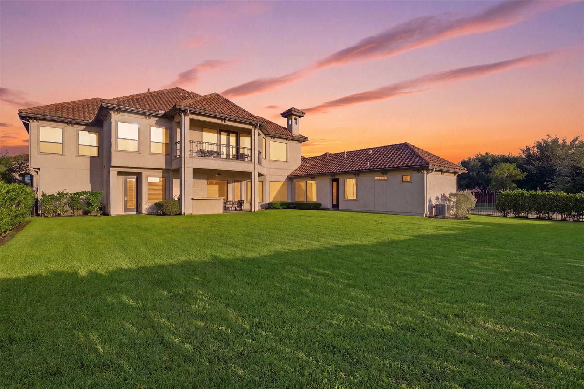 Rear view showing the home's stucco exterior, barrel tile roof, dual covered verandas, and expansive lawn space.
