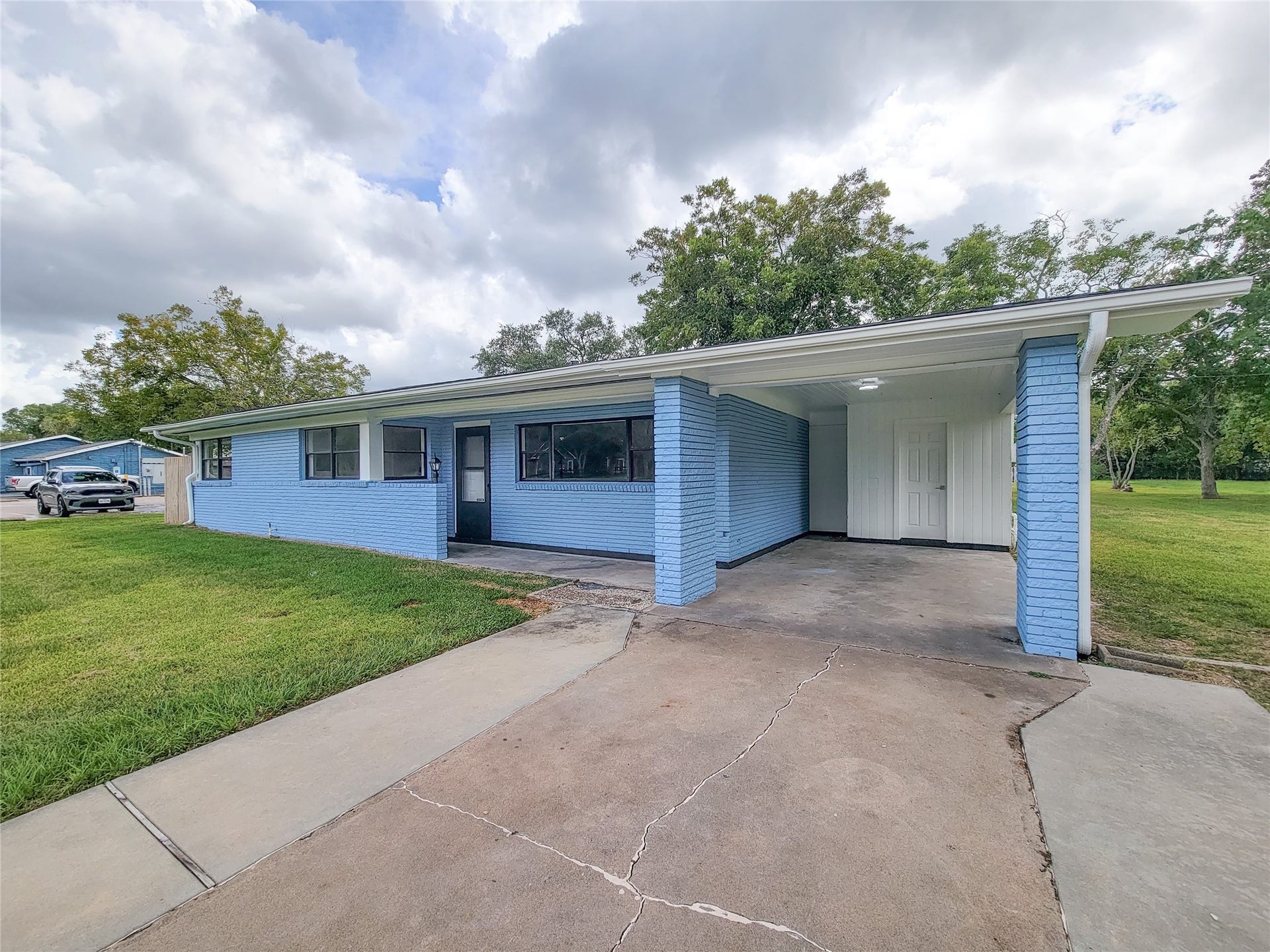 2605 W Dumble Street, Alvin, TX 77511 Charming blue brick home with a spacious carport, nestled on a lush green lawn and surrounded by mature trees, offering serene and picturesque living.
