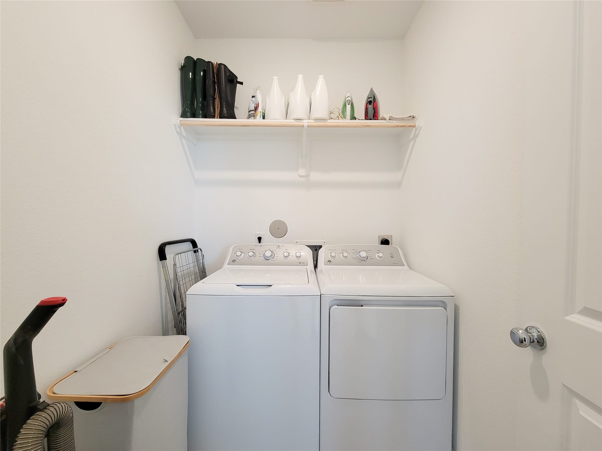2719 Forest Retreat Lane, Missouri City, TX 77489 Compact laundry room with a washer and dryer set. It includes a shelf above for storage, holding items like detergent and an iron. The space is well-organized and has a clean, white finish.