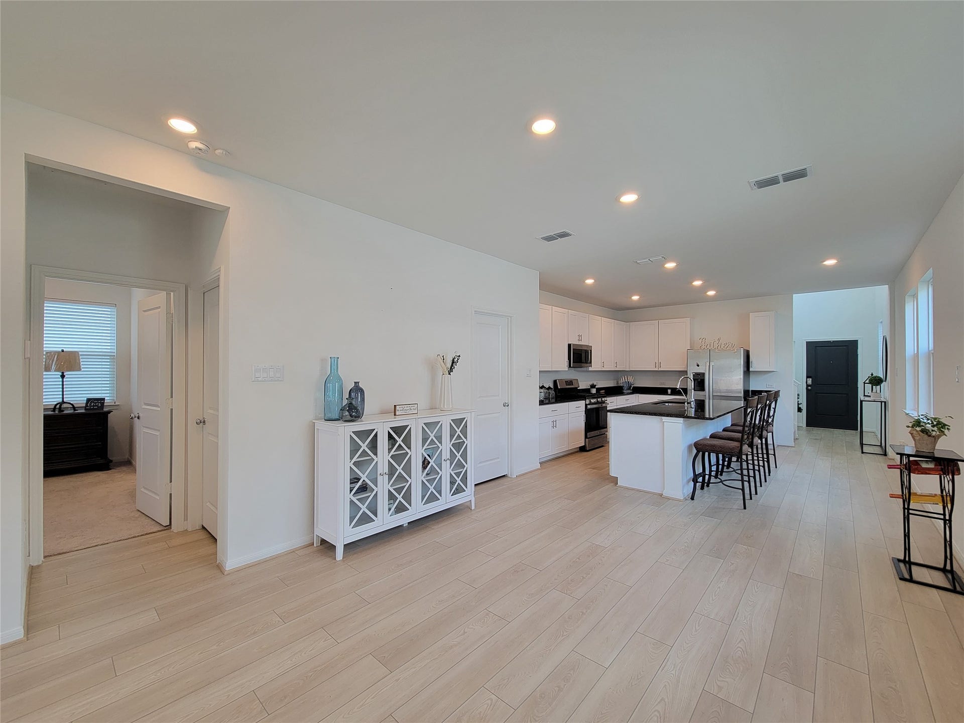 2719 Forest Retreat Lane, Missouri City, TX 77489 Bright, open-concept kitchen and living area with light wood flooring. The kitchen features modern white cabinetry, stainless steel appliances, and a central island with seating. A doorway leads to a cozy bedroom, and the space is well-lit with recessed lighting.