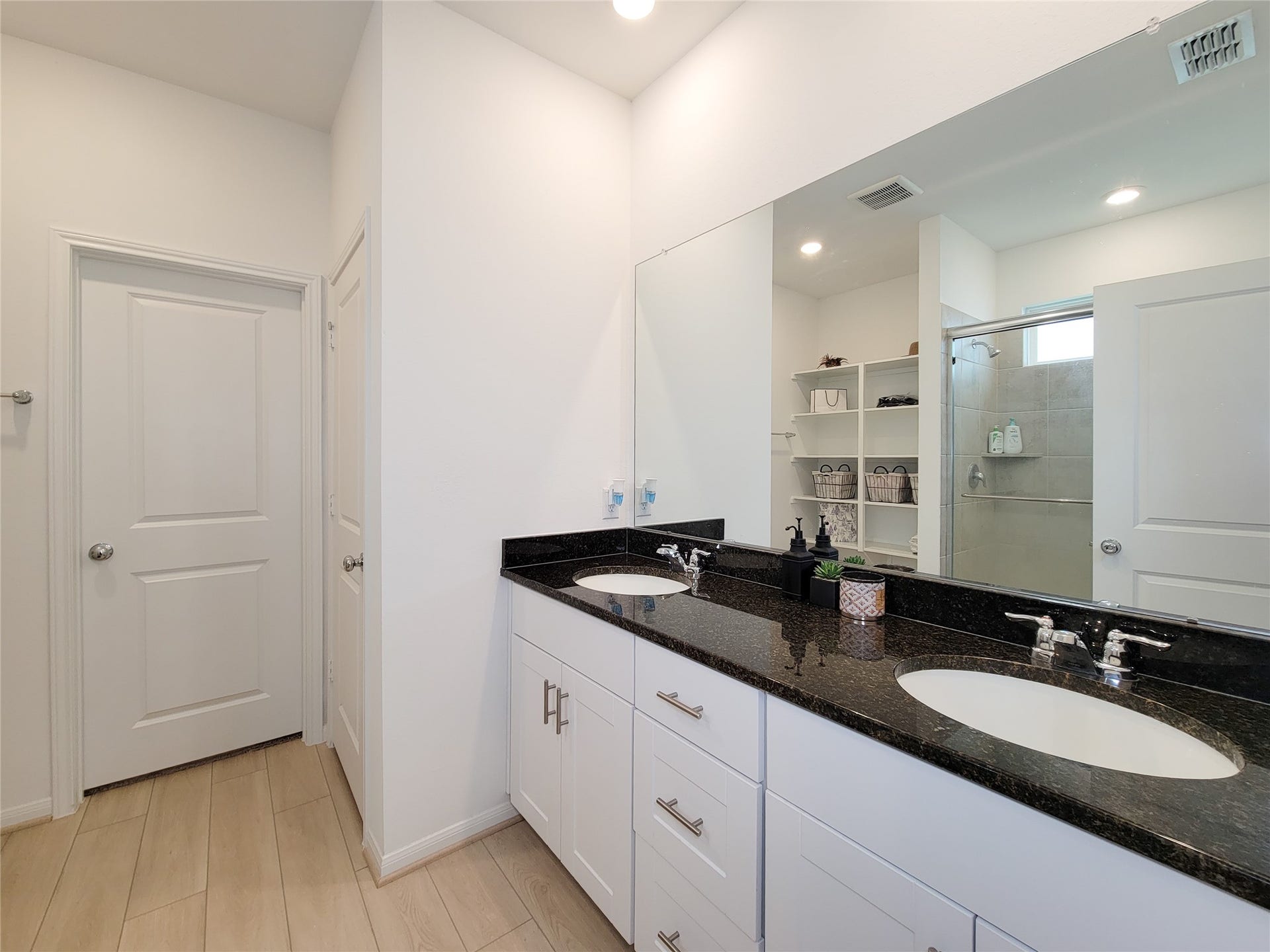 2719 Forest Retreat Lane, Missouri City, TX 77489 Modern bathroom with dual sinks set in a sleek black countertop, white cabinetry, and a large mirror. The room features light wood flooring, recessed lighting, and a walk-in shower with glass doors. Ample storage space with open shelving enhances functionality.