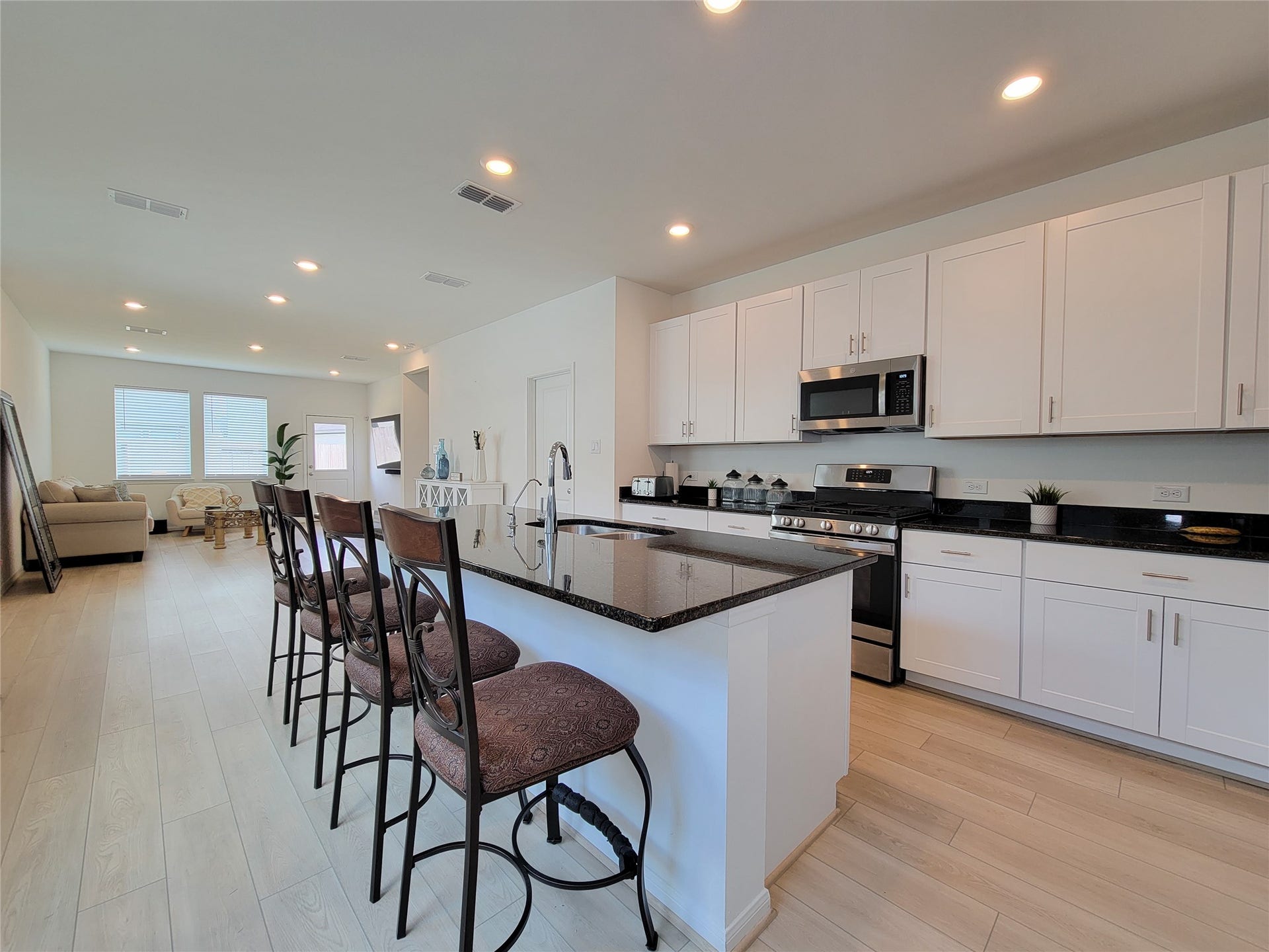 2719 Forest Retreat Lane, Missouri City, TX 77489 Modern, open-concept kitchen and living area. The kitchen features sleek white cabinetry, a large island with bar seating, and stainless steel appliances. The space is well-lit with recessed lighting, and the light wood flooring adds a warm touch. The living area in the background offers ample natural light from large windows.