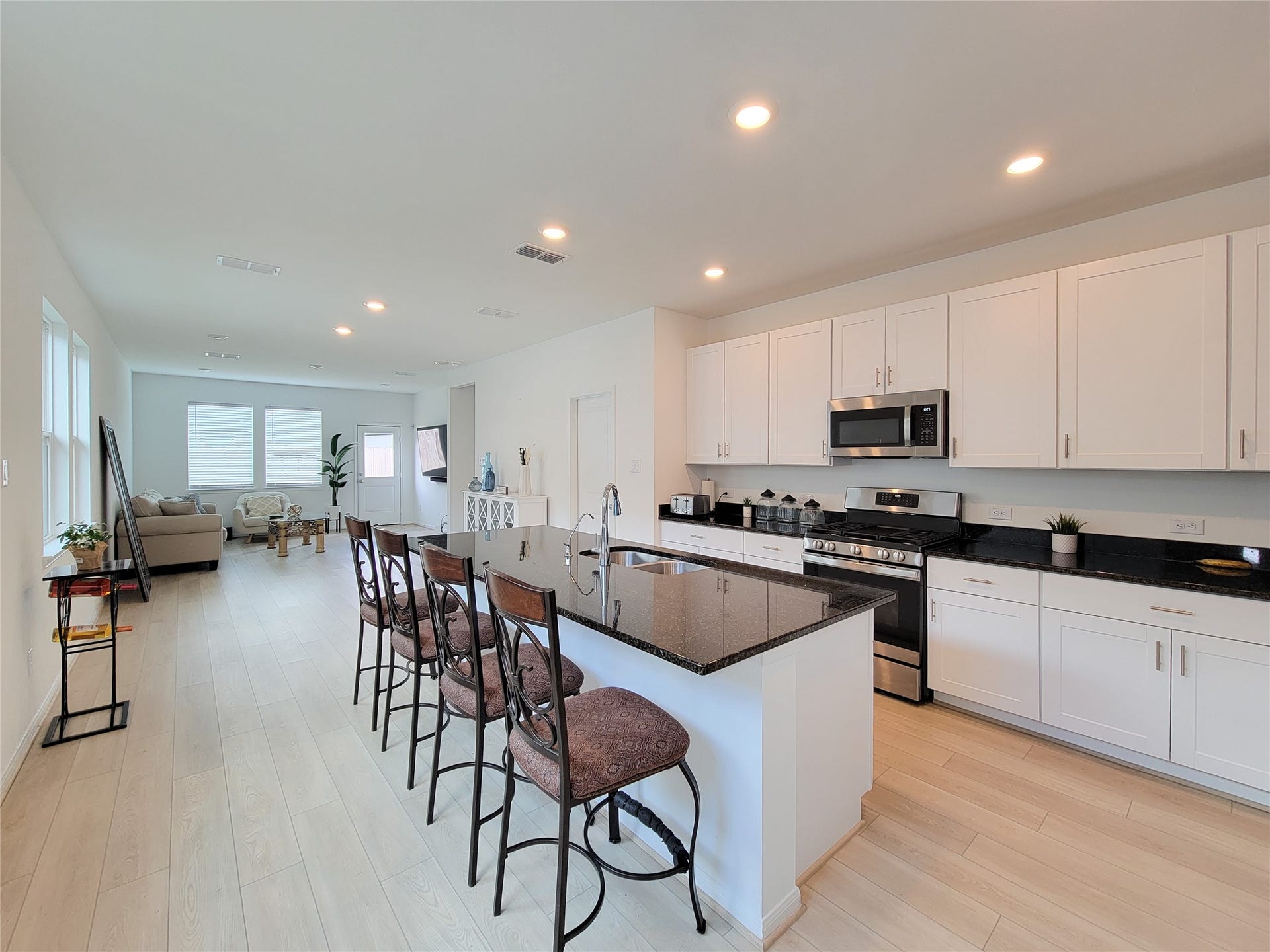 2719 Forest Retreat Lane, Missouri City, TX 77489 The kitchen features modern white cabinets, a sleek black granite countertop, and a central island with bar seating. The space is well-lit with recessed lighting, and the adjacent living area offers ample natural light, providing a warm and inviting atmosphere.