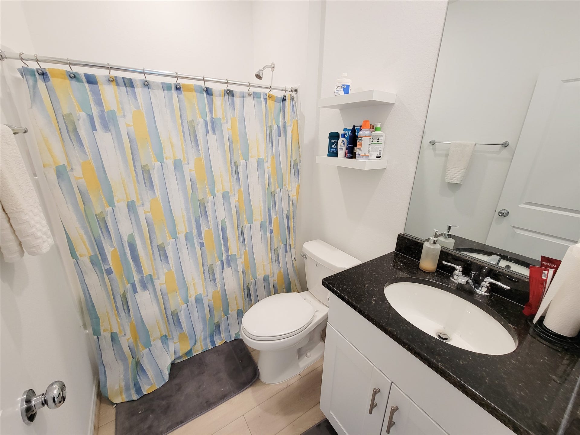 2719 Forest Retreat Lane, Missouri City, TX 77489 This bathroom features a modern design with a black granite countertop, white vanity, and a colorful shower curtain. It includes a standard toilet, shelves for storage, and a large mirror. The space is well-lit and has a clean, fresh appearance.