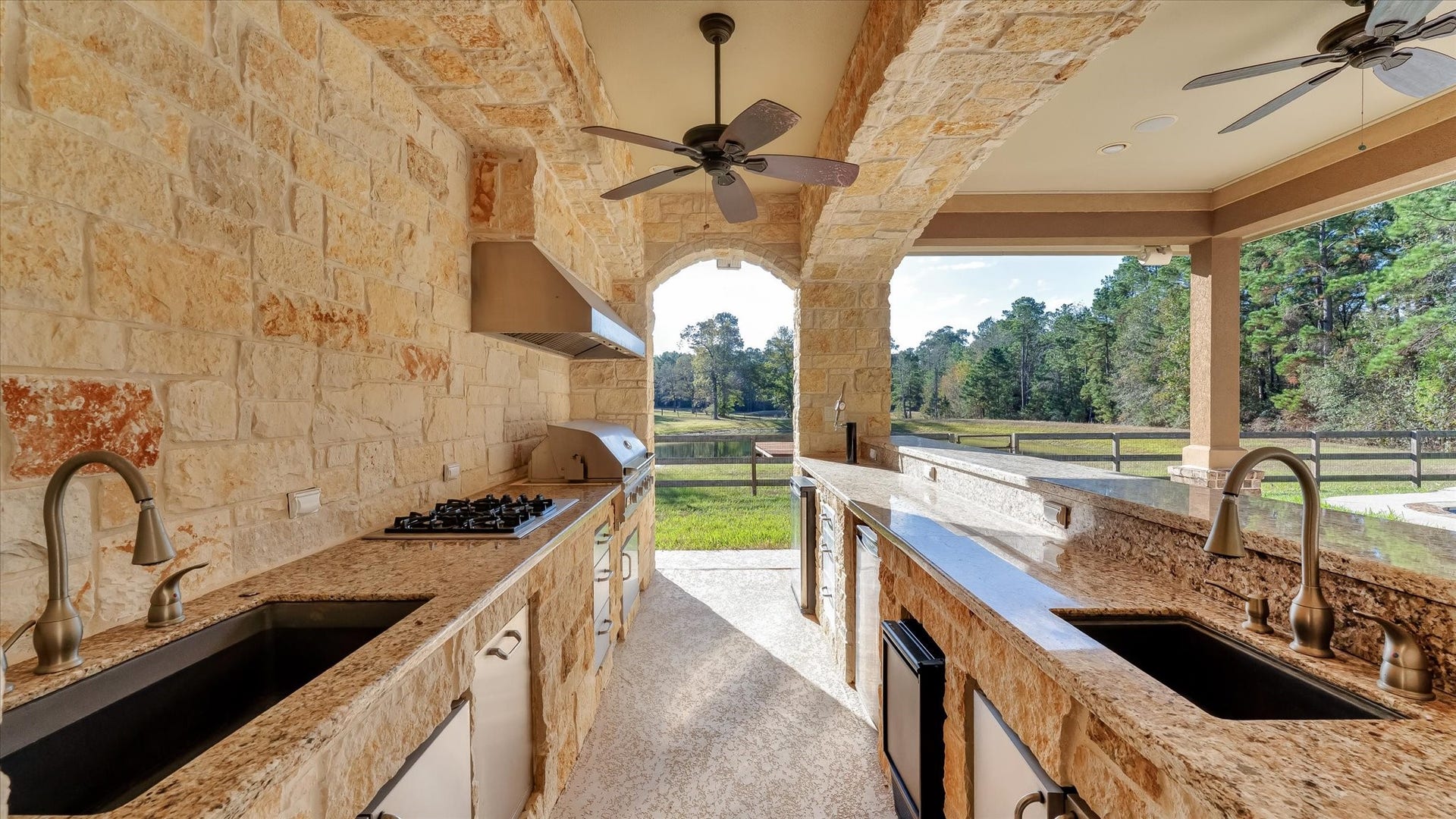 8071 Stacy Diane Drive, Montgomery, TX 77316 This photo showcases a spacious outdoor kitchen with stone walls and countertops, equipped with a built-in cooktop and dual sinks. Ceiling fans provide comfort, and the space opens up to a scenic view of greenery and trees, perfect for entertaining and enjoying nature.