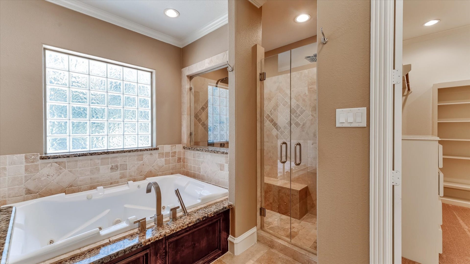 8071 Stacy Diane Drive, Montgomery, TX 77316 This bathroom features a luxurious jetted tub with granite countertops, a spacious glass-enclosed shower, and elegant tile work. Natural light streams through a large glass block window, enhancing the warm, inviting atmosphere.