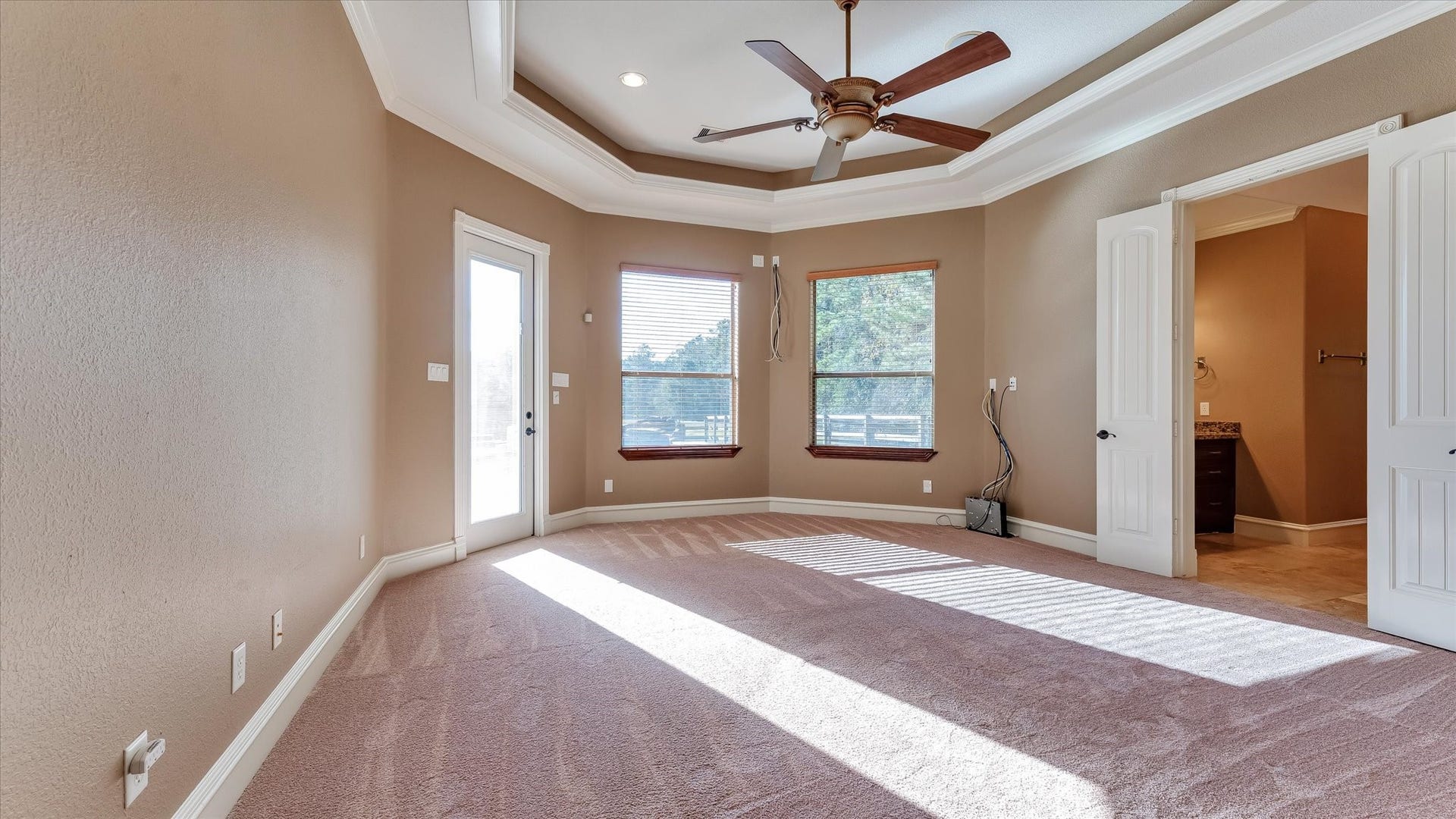 8071 Stacy Diane Drive, Montgomery, TX 77316 This spacious bedroom features a tray ceiling with a ceiling fan, large windows for natural light, and a doorway leading to a patio. It has warm beige walls, plush carpeting, and connects to a bathroom through double doors.