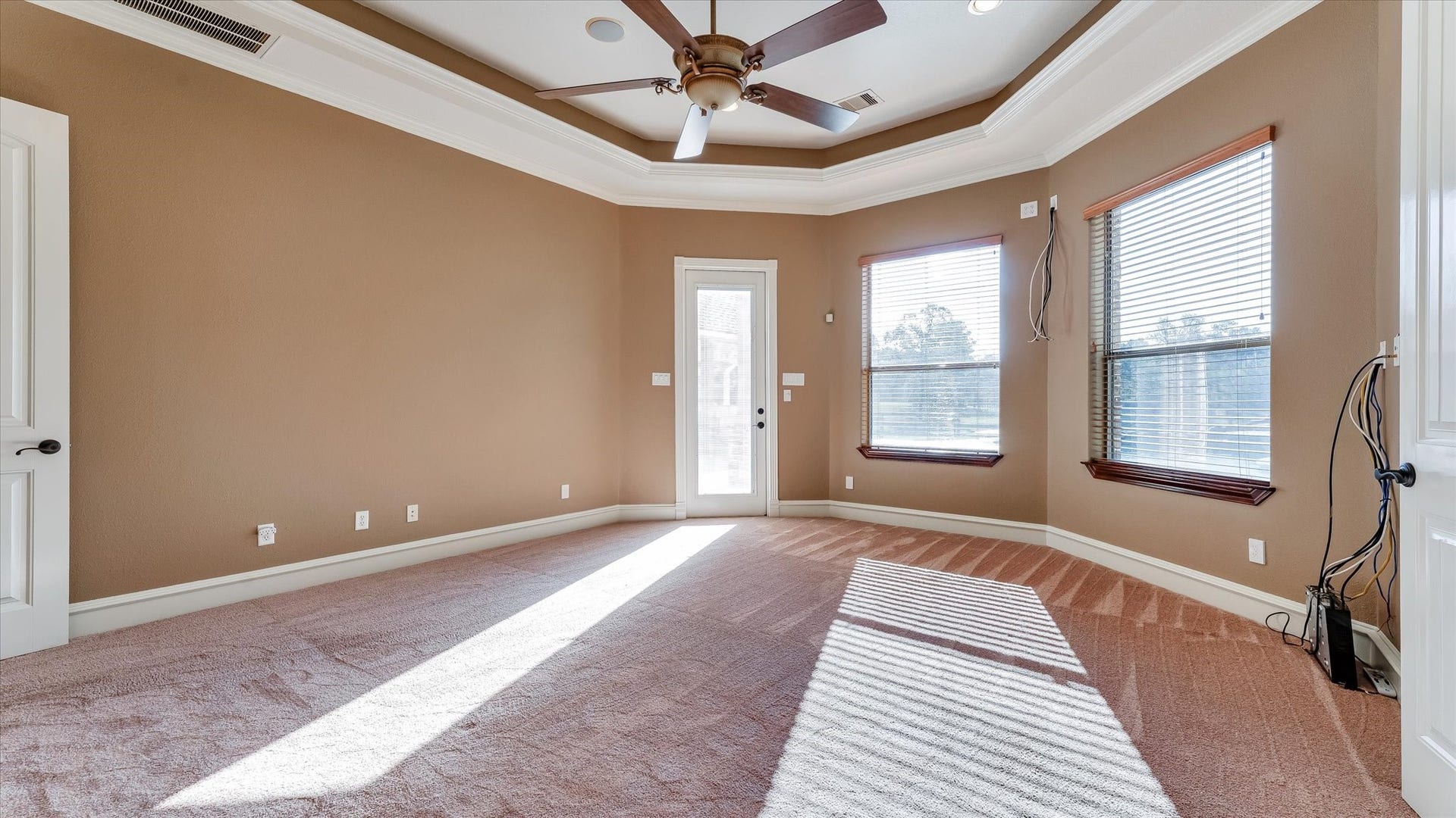 8071 Stacy Diane Drive, Montgomery, TX 77316 This spacious room features a tray ceiling with a ceiling fan, neutral walls, and ample natural light from large windows. It has a cozy carpeted floor and a door leading outside, ideal for a comfortable and bright living space.