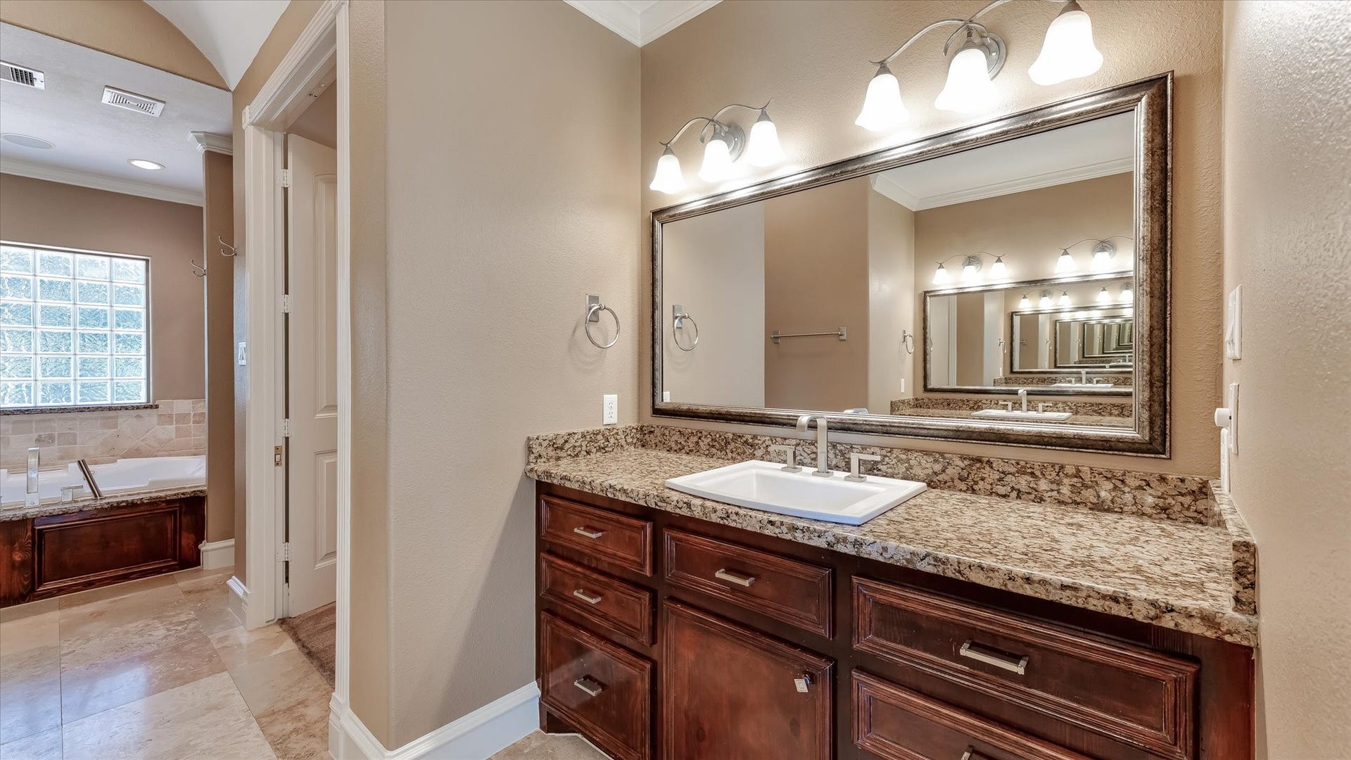 8071 Stacy Diane Drive, Montgomery, TX 77316 This bathroom features a large mirror with modern lighting above a granite countertop and wooden cabinetry. To the side, there's a spacious tub area with natural light from a glass block window. The space is elegant and inviting.