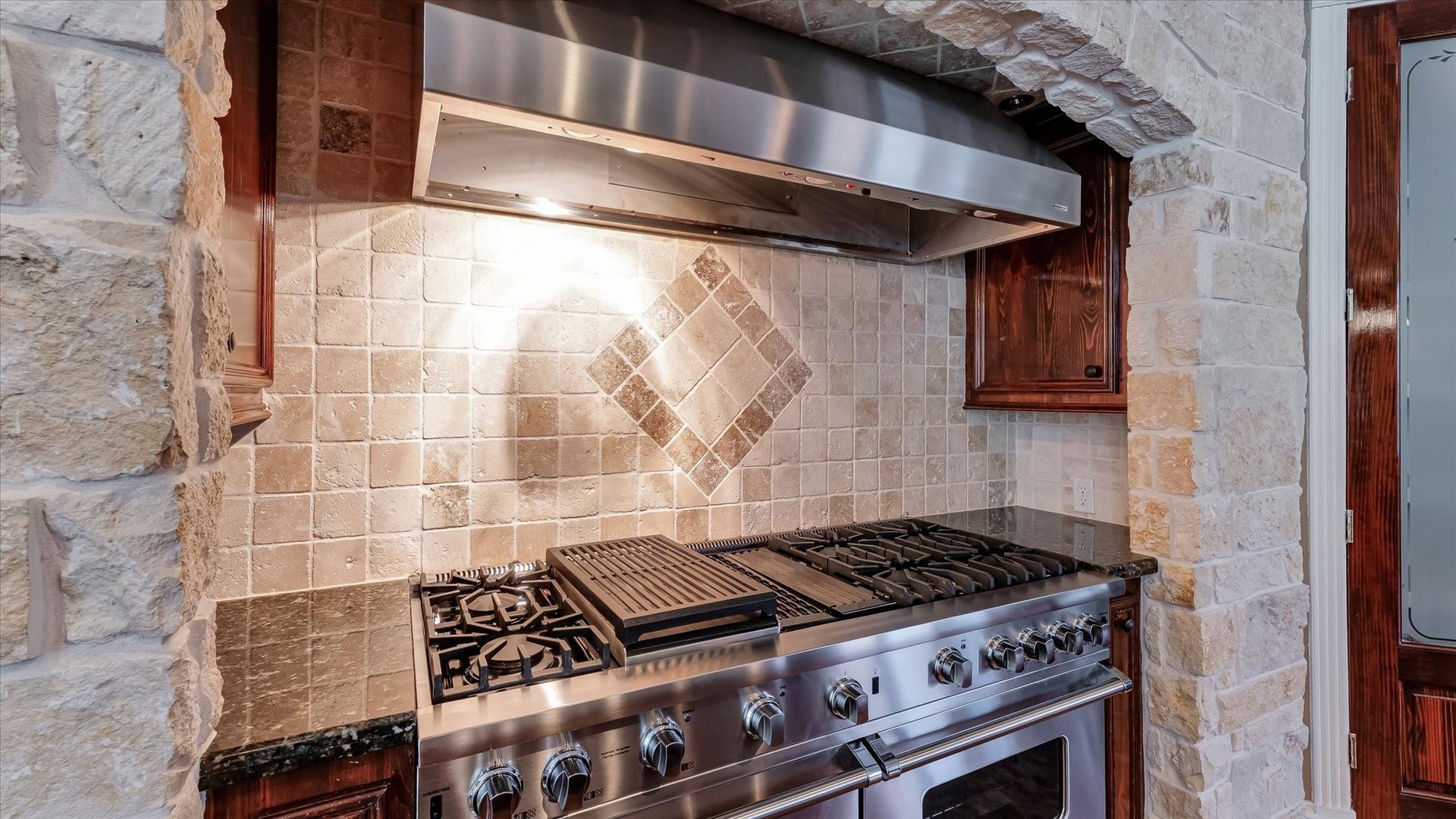 8071 Stacy Diane Drive, Montgomery, TX 77316 This kitchen features a high-end stainless steel gas range with multiple burners and a griddle, set against a stylish tiled backsplash with a diamond accent. It's framed by rustic stone walls and dark wood cabinetry, creating a warm, inviting cooking space.