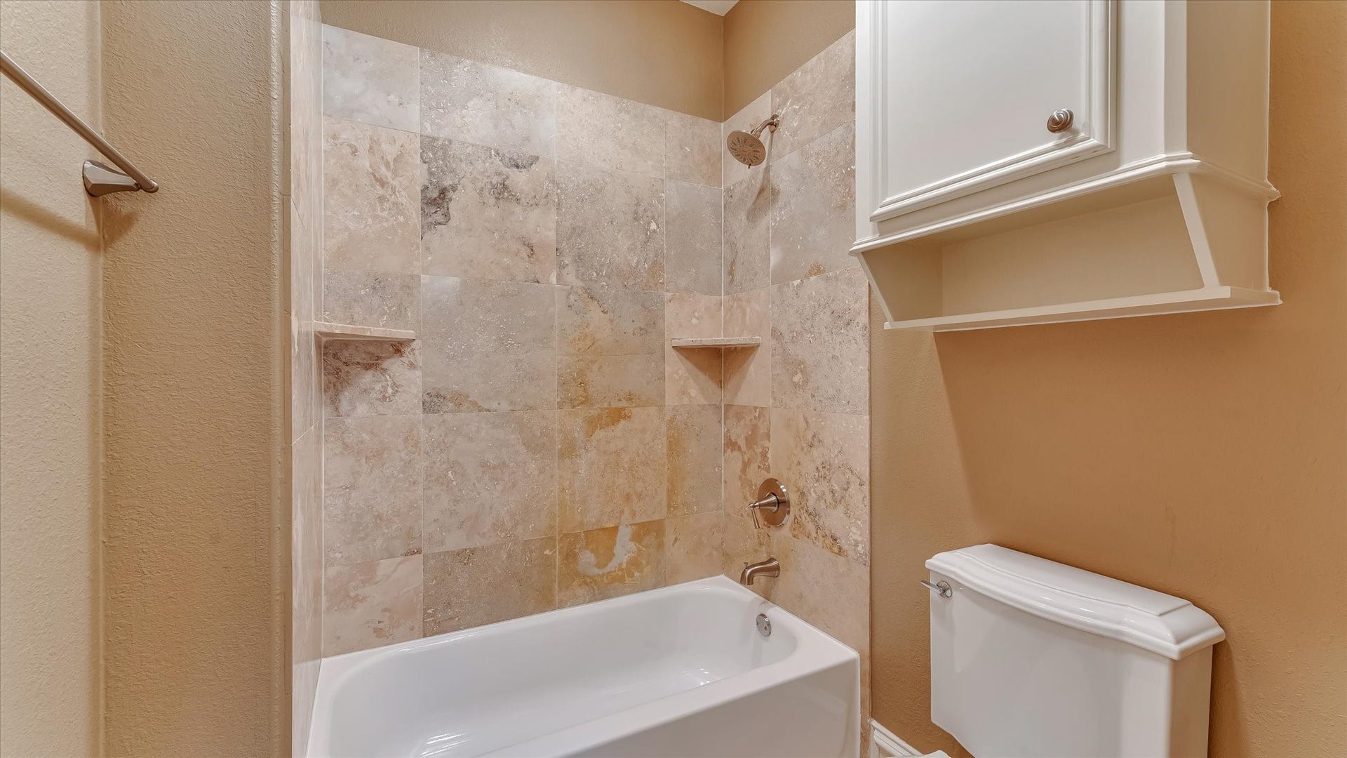 8071 Stacy Diane Drive, Montgomery, TX 77316 This bathroom features a classic tub-shower combo with elegant stone tile walls. It has a modern showerhead and built-in corner shelves. The white cabinetry offers additional storage above the toilet, and the neutral walls create a warm, inviting atmosphere.