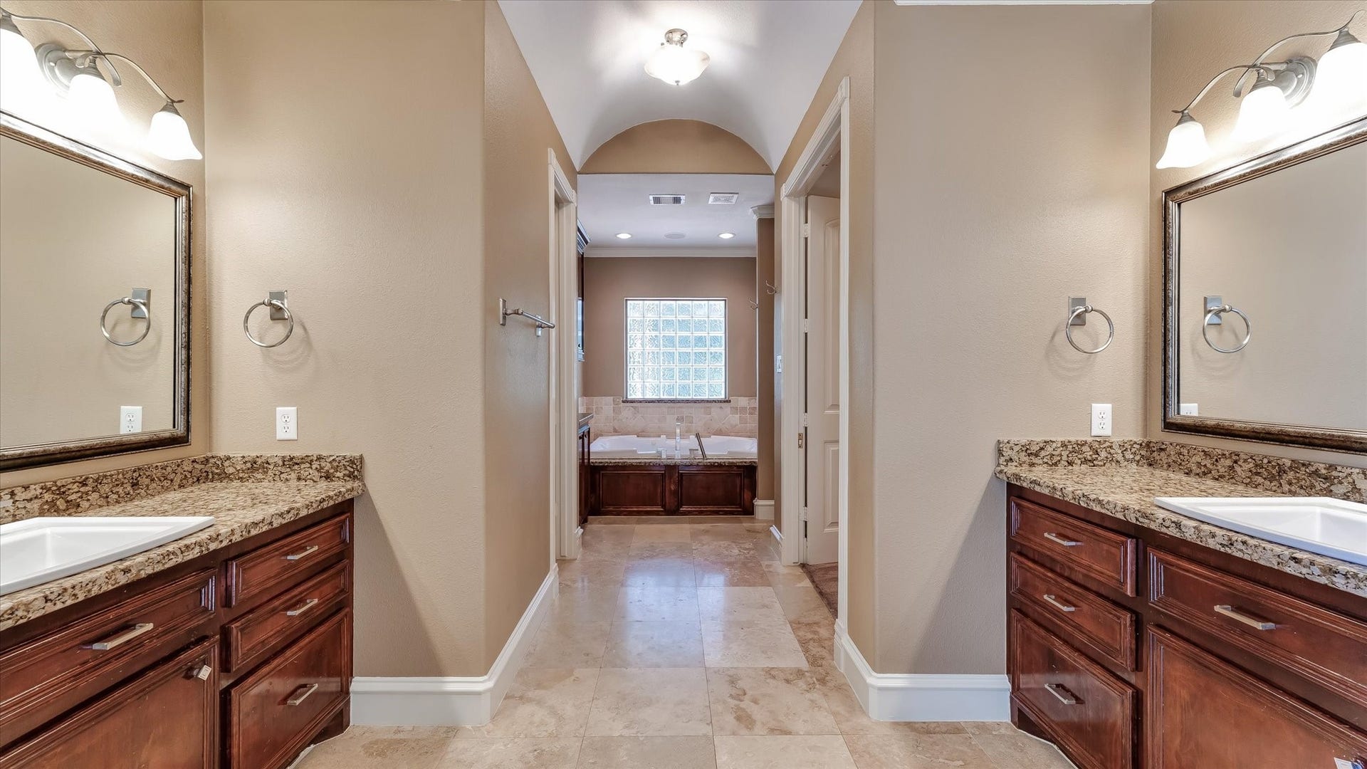 8071 Stacy Diane Drive, Montgomery, TX 77316 This photo showcases a spacious bathroom with dual sinks and granite countertops, rich wooden cabinetry, and a neutral color palette. The room features a central pathway leading to a large bathtub under a window, offering a relaxing atmosphere.