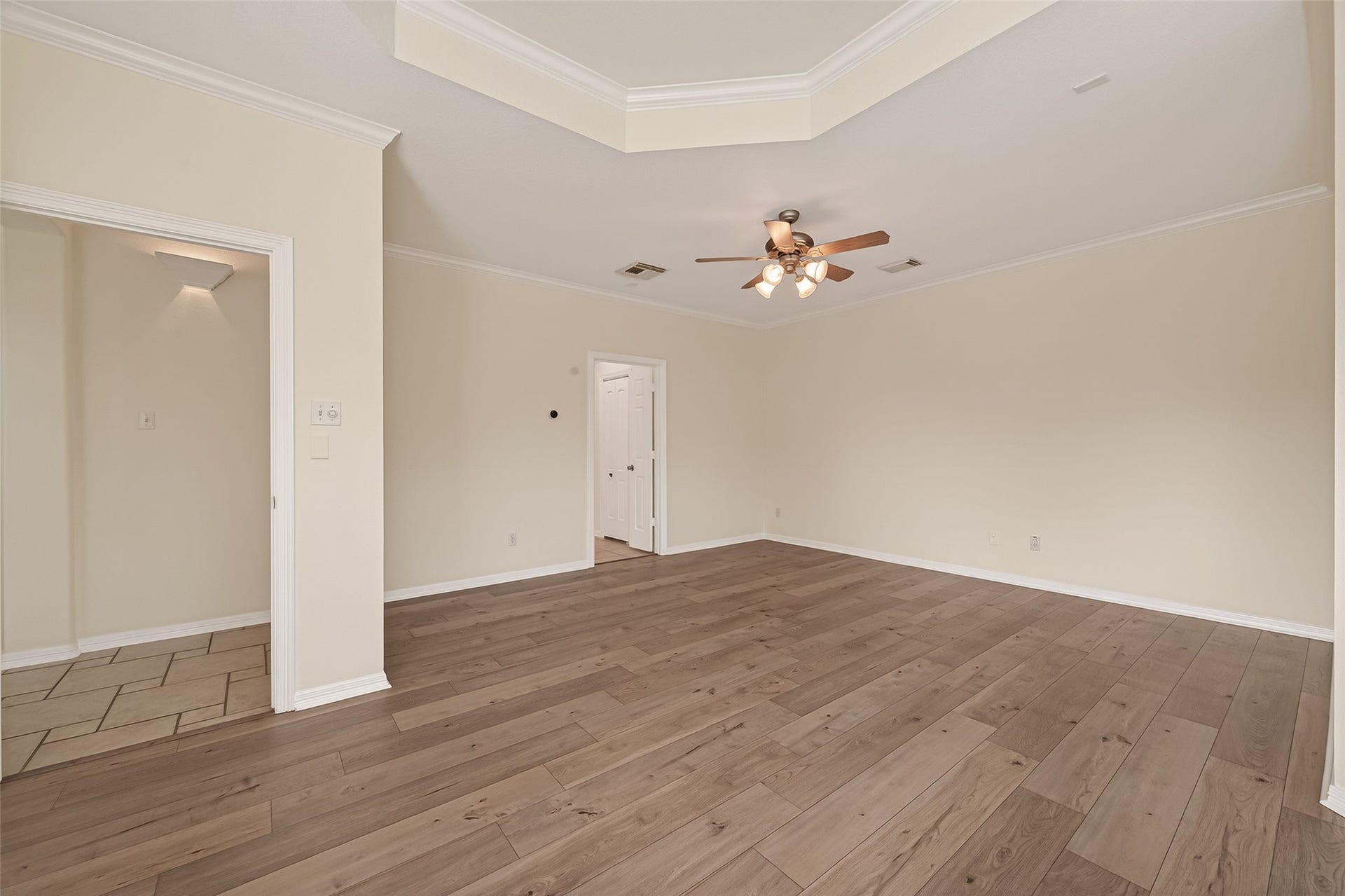 10106 Lost Hollow Lane, Missouri City, TX 77459 This spacious primary bedroom pfeatures elegant wood flooring, a ceiling fan with lights, and neutral wall colors. The room is well-lit and includes a decorative tray ceiling, offering a stylish and inviting atmosphere.