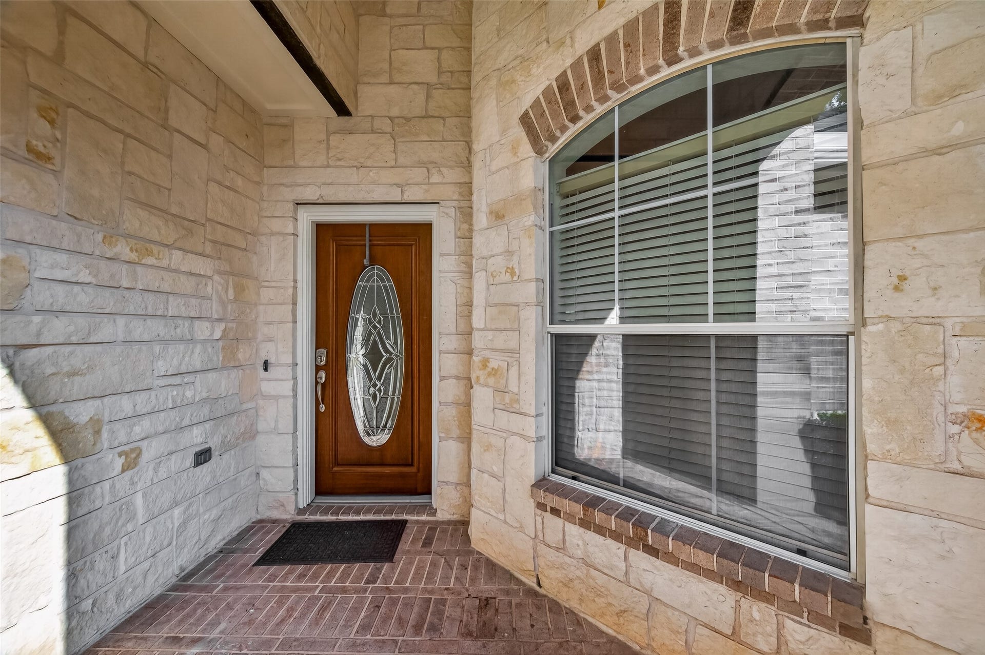 10106 Lost Hollow Lane, Missouri City, TX 77459 welcoming entryway featuring a wooden door with an ornate glass panel, surrounded by stone walls and a large window with blinds. The space is accented by a brick-textured floor, creating a charming and inviting first impression for potential homebuyers.