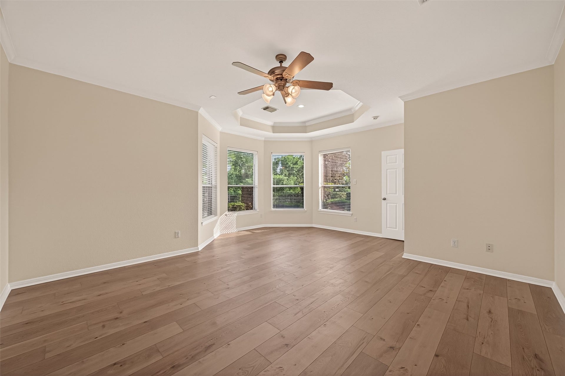 10106 Lost Hollow Lane, Missouri City, TX 77459 Spacious primary bedroom with warm wood flooring, neutral walls, and a tray ceiling with a fan. Large windows provide natural light and a view of greenery, creating a bright and inviting space.
