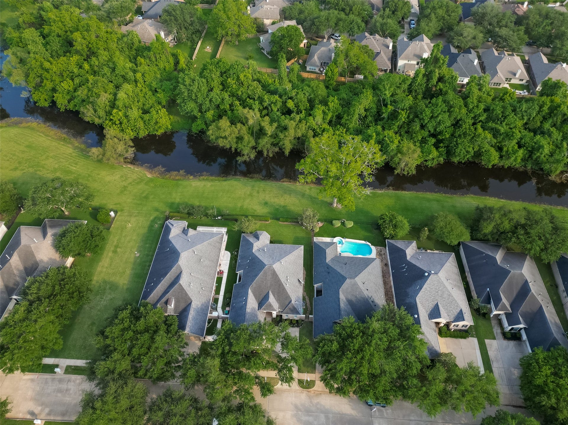 10106 Lost Hollow Lane, Missouri City, TX 77459 This aerial photo showcases a row of suburban homes with spacious yards, one featuring a swimming pool. The properties back onto a lush, wooded area with a small waterway, offering privacy and natural views. The neighborhood appears quiet and well-maintained.