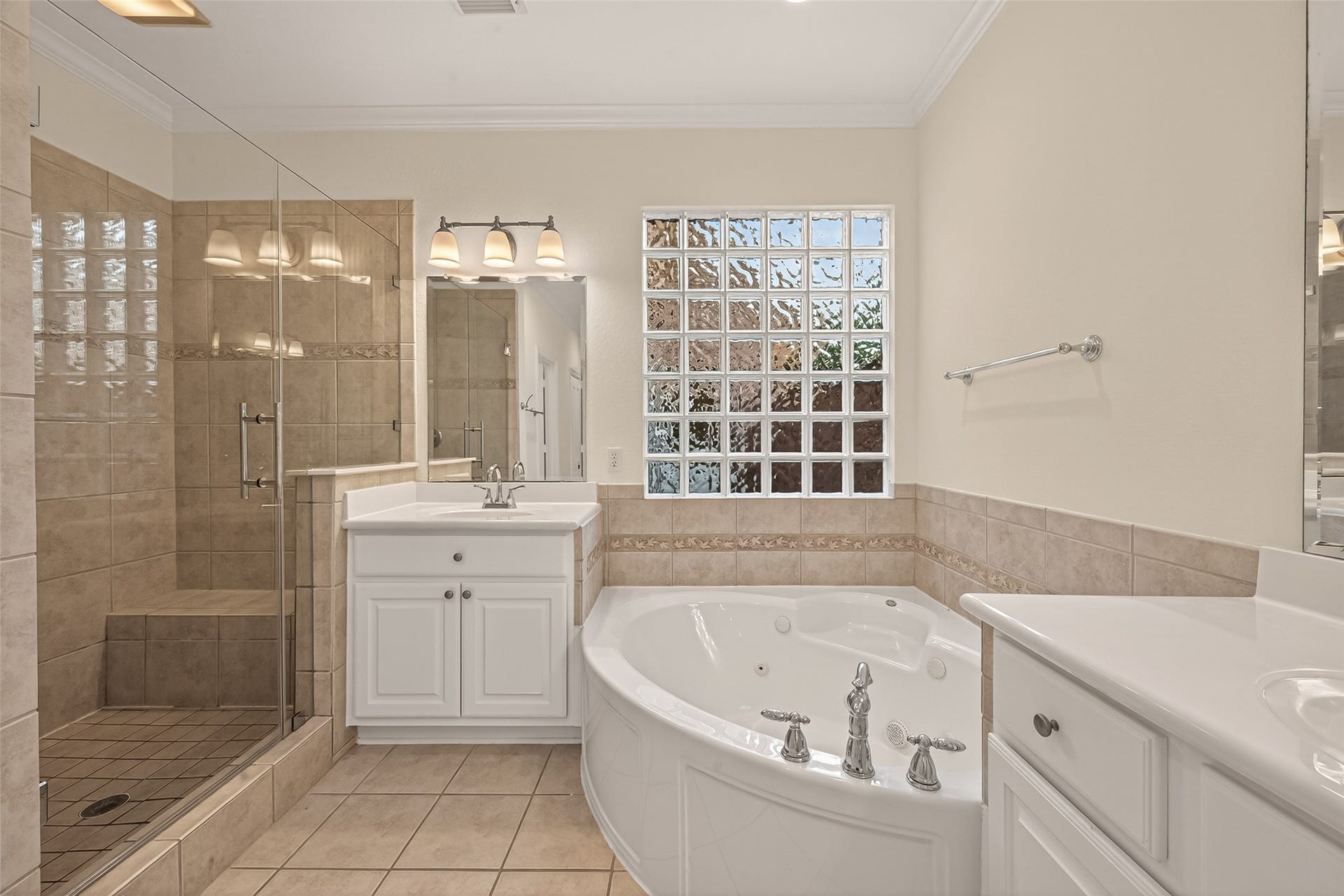 10106 Lost Hollow Lane, Missouri City, TX 77459 This bathroom features a spacious walk-in shower with glass doors, a luxurious corner jetted tub, dual sinks with white cabinetry, and a large glass block window for natural light. The neutral tile and clean design create a relaxing atmosphere.