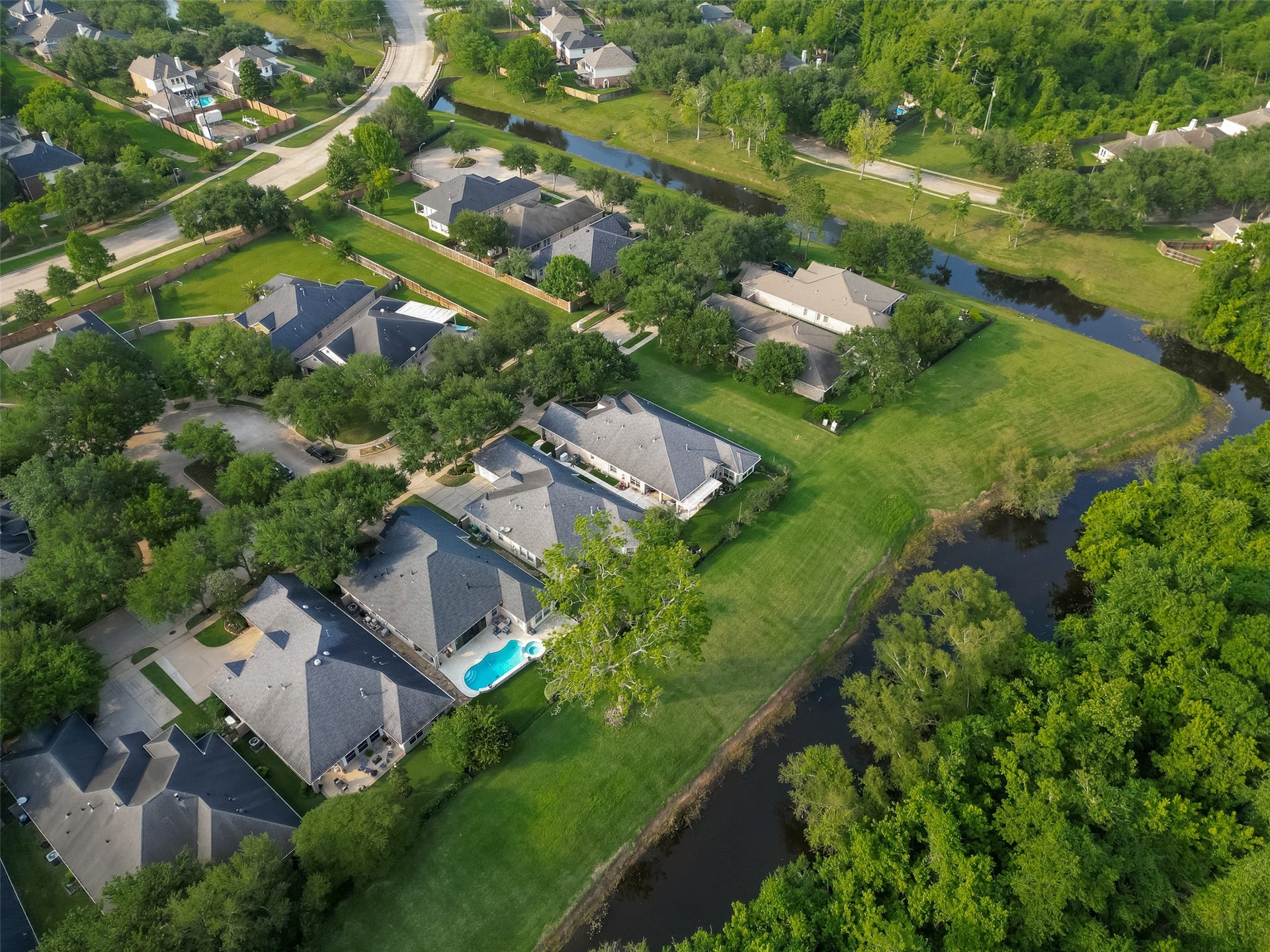 10106 Lost Hollow Lane, Missouri City, TX 77459 This aerial view showcases a serene neighborhood with spacious homes surrounded by lush greenery. The properties have well-maintained lawns, and one features a private pool. A small body of water and wooded areas add to the natural charm, offering a peaceful environment.
