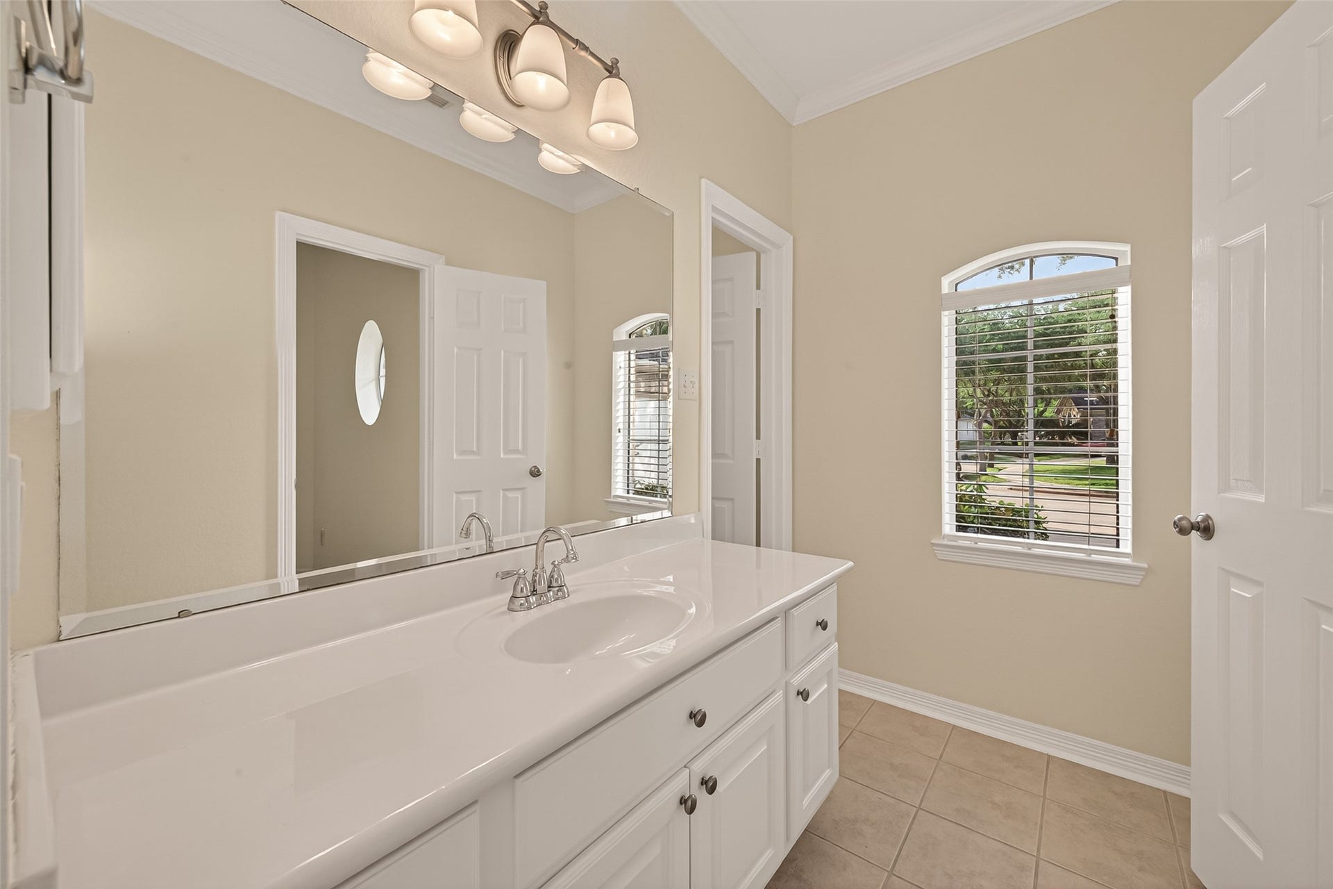10106 Lost Hollow Lane, Missouri City, TX 77459 This bathroom features a spacious vanity with dual sinks, elegant lighting fixtures, and a large mirror. It has a window with blinds for natural light and privacy, complemented by neutral tile flooring and walls.