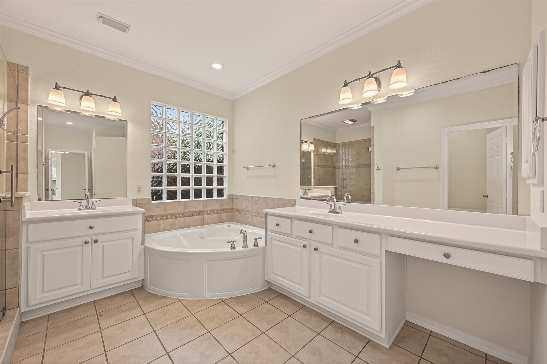 10106 Lost Hollow Lane, Missouri City, TX 77459 This primary bathroom features a spacious layout with dual vanities, a large mirror, and a luxurious corner bathtub. It includes a glass block window for natural light, a separate shower, and ample cabinet storage. The neutral tile flooring and soft lighting create a relaxing ambiance.
