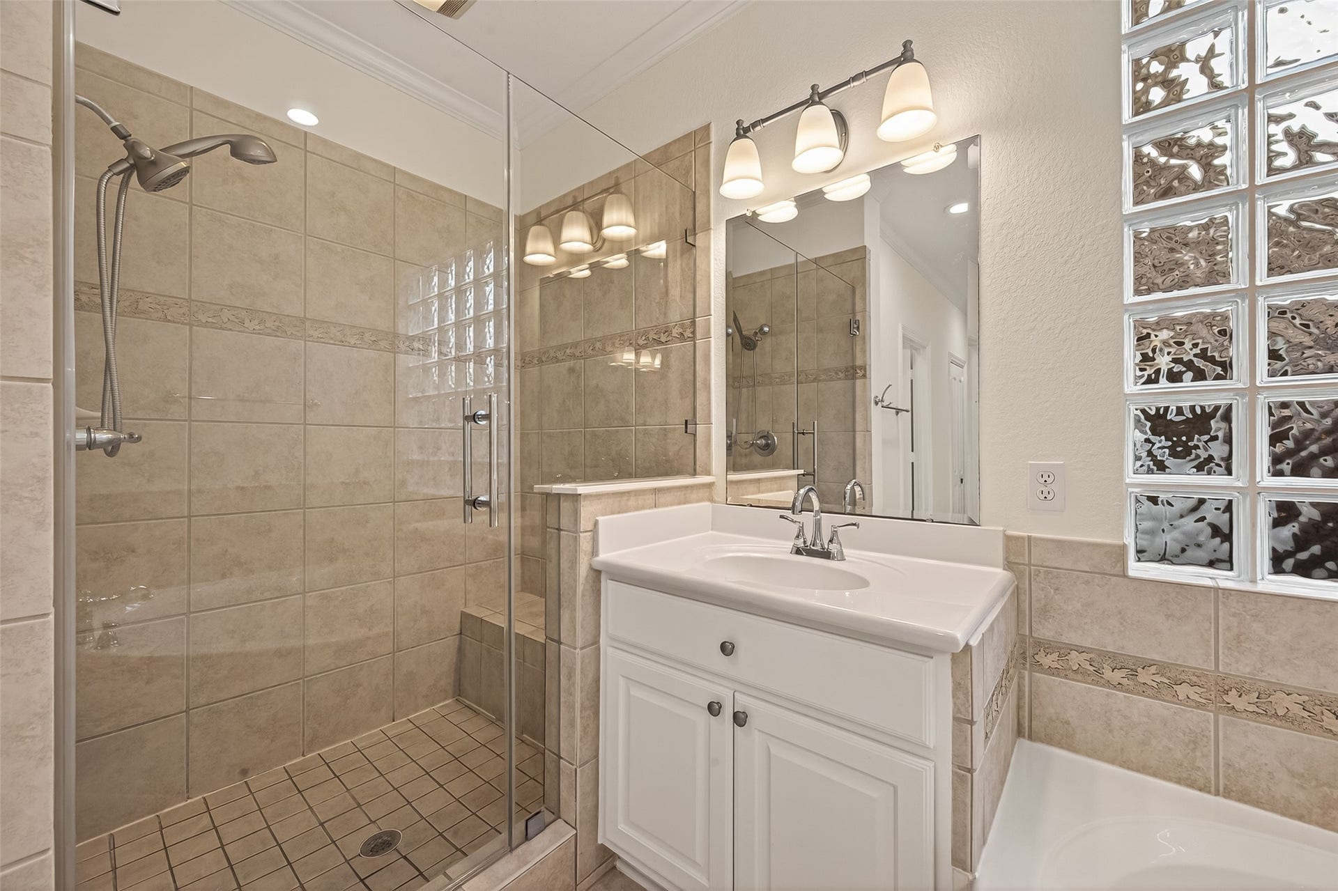 10106 Lost Hollow Lane, Missouri City, TX 77459 This bathroom features a modern glass-enclosed shower with stylish beige tiles, a sleek white vanity with a large mirror, and elegant lighting fixtures. The room is brightened by glass block windows, adding a touch of sophistication.