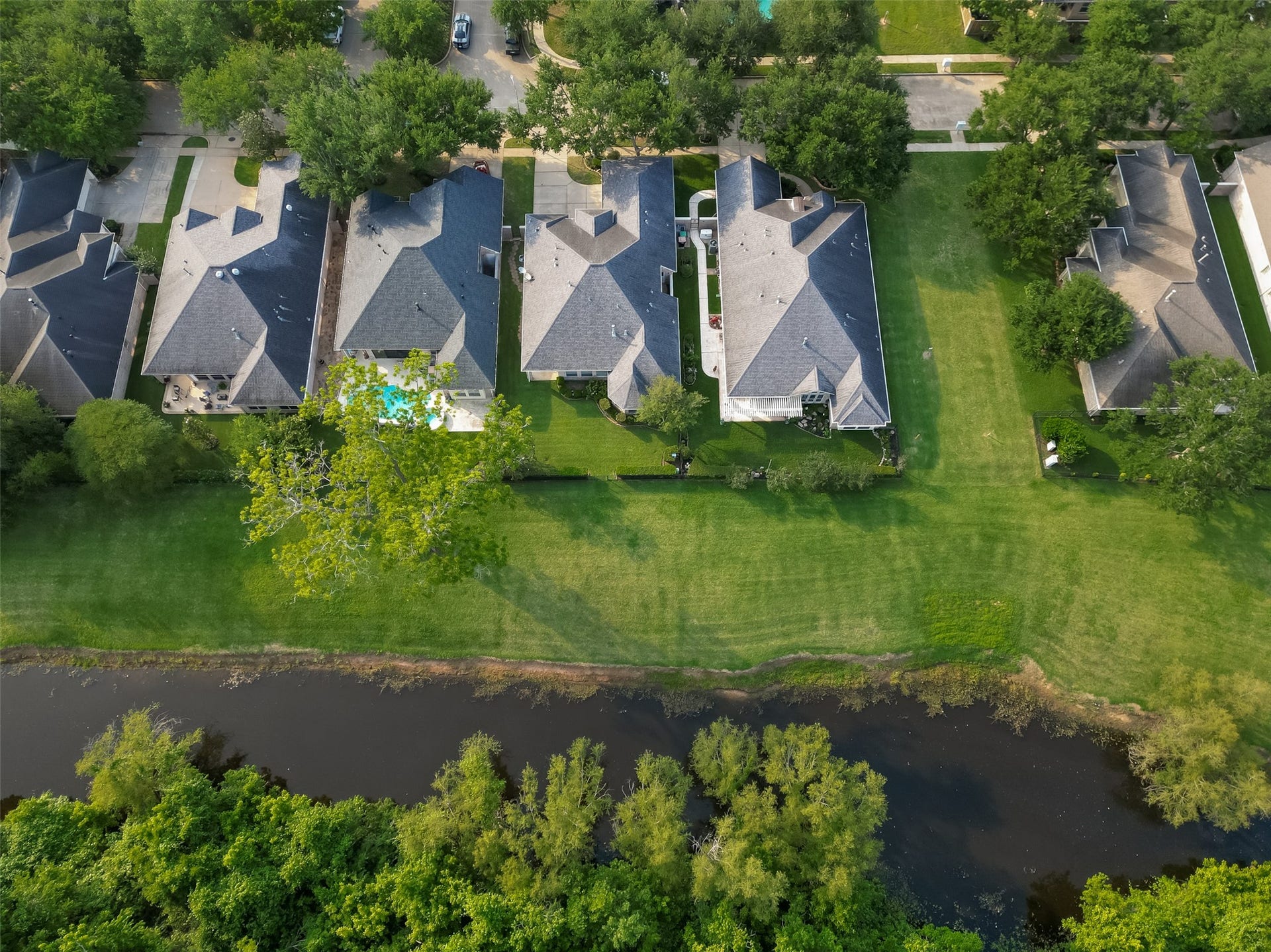 10106 Lost Hollow Lane, Missouri City, TX 77459 This aerial view showcases a row of well-maintained suburban homes with spacious backyards, some featuring pools. The properties back onto a lush green area with a waterway, offering a scenic and serene environment.