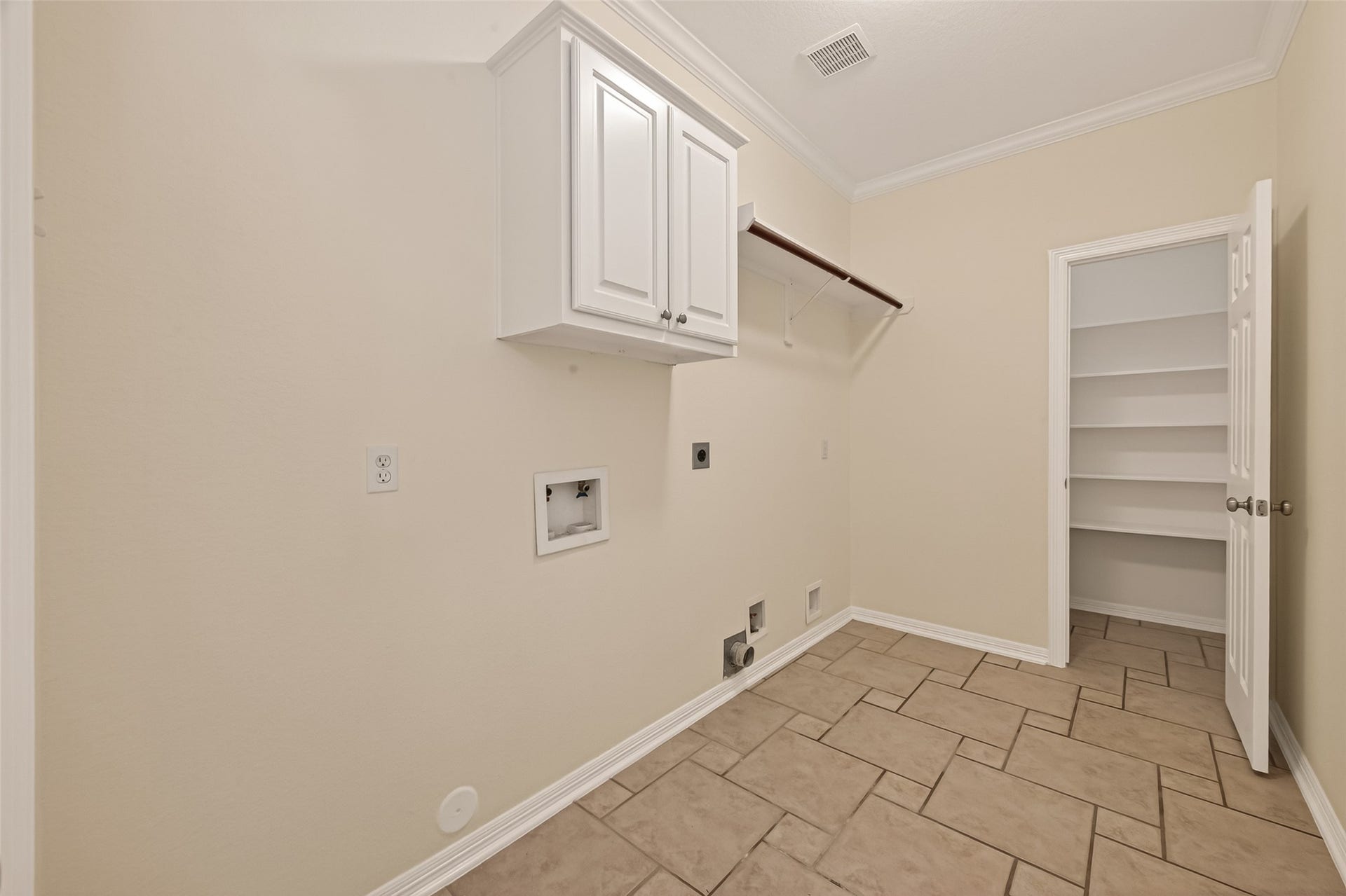 10106 Lost Hollow Lane, Missouri City, TX 77459 This photo shows a spacious laundry room with beige tile flooring, a wall-mounted cabinet for storage, and a rod for hanging clothes. It features hookups for a washer and dryer, and an open door leads to additional shelving space.