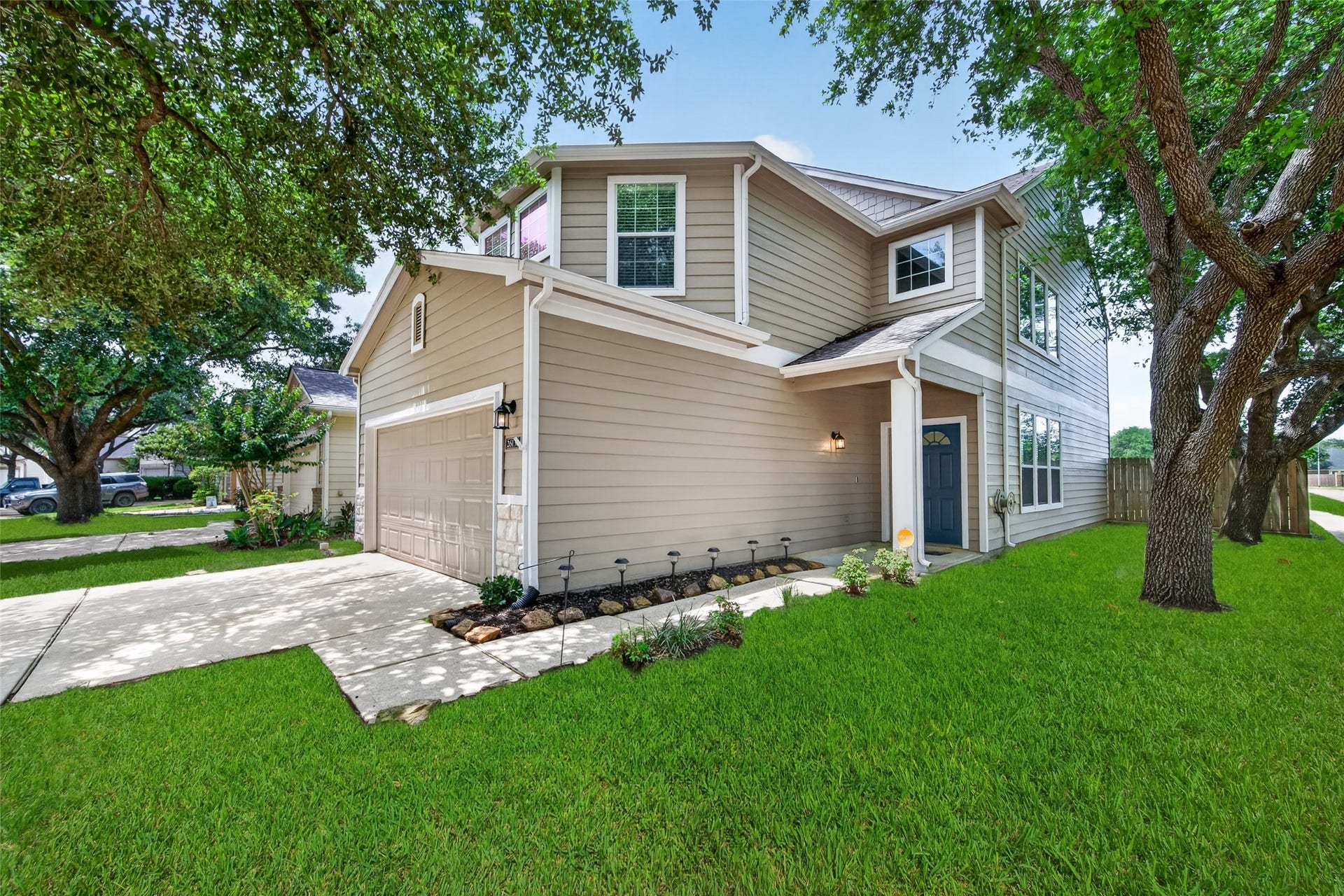26130 Ripley Hills Drive, Richmond, TX 77406 This photo shows a charming two-story home with beige siding and white trim. It features a double garage, a well-maintained front lawn, and mature trees providing shade. The entrance has a welcoming porch with a blue door, giving the home a cozy and inviting feel.