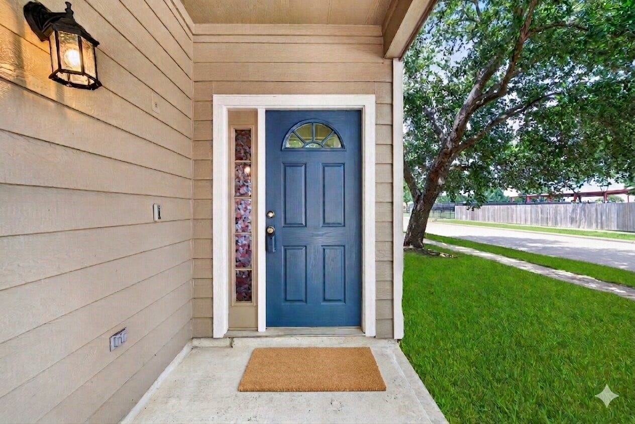 26130 Ripley Hills Drive, Richmond, TX 77406 Charming front entry with a blue door framed by a side window, set against neutral siding. A welcoming light fixture and doormat add warmth, while the lush green lawn and tree provide a serene outdoor view.