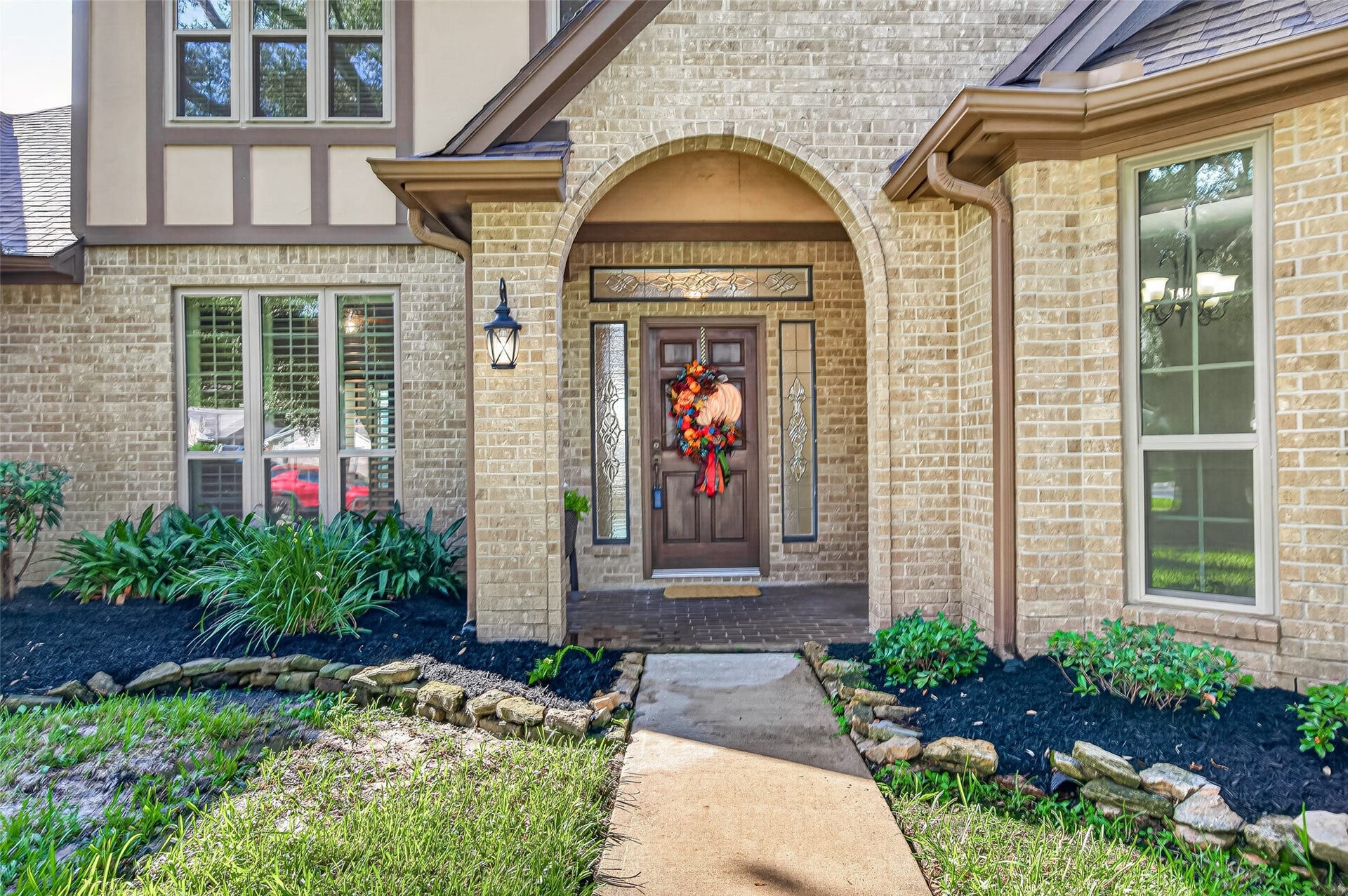 A CHARMING ENTRY THAT SAYS, "COME ON IN!" Follow the walkway past beautifully landscaped flowerbeds to the inviting covered front porch. A graceful brick archway frames the handsome wood front door with decorative leaded-glass sidelights & transom. Imagine greeting friends beneath the covered entry, decorating for your favorite holidays or enjoying a gentle rain from the shelter of the porch. A traditional coach light adds a welcoming glow after sunset. What a lovely place to come home to.