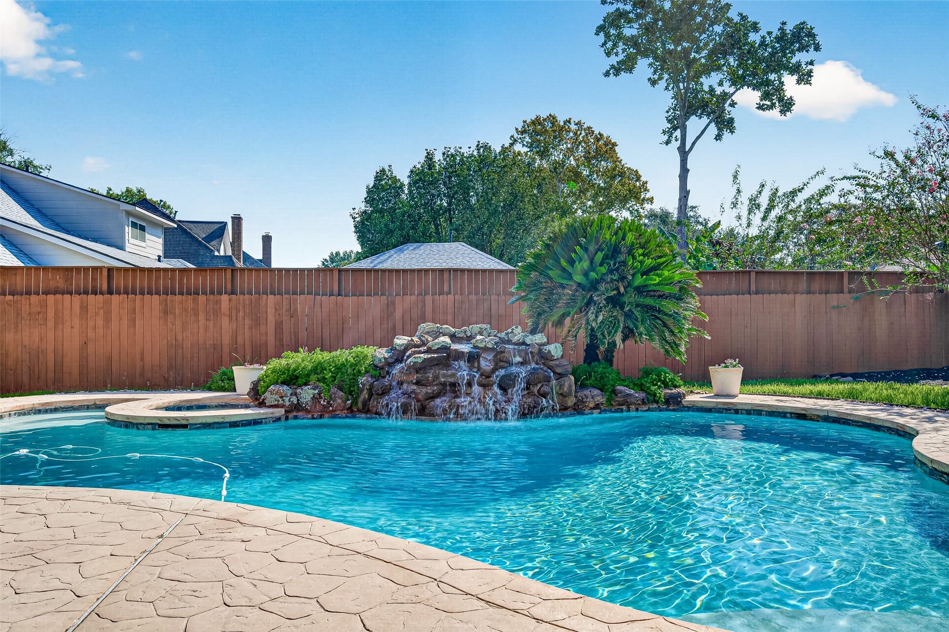 LET THE SOUND OF THE WATERFALL WASH THE DAY AWAY! Imagine relaxing poolside as water cascades over the beautiful rock waterfall, creating a soothing backdrop for a peaceful afternoon. Tropical palms, lush landscaping & sparkling blue water give this backyard a resort-like atmosphere. Whether you're enjoying a quiet morning outdoors or unwinding after a busy day, this inviting retreat offers a wonderful place to escape the everyday hustle & bustlewithout ever leaving home!