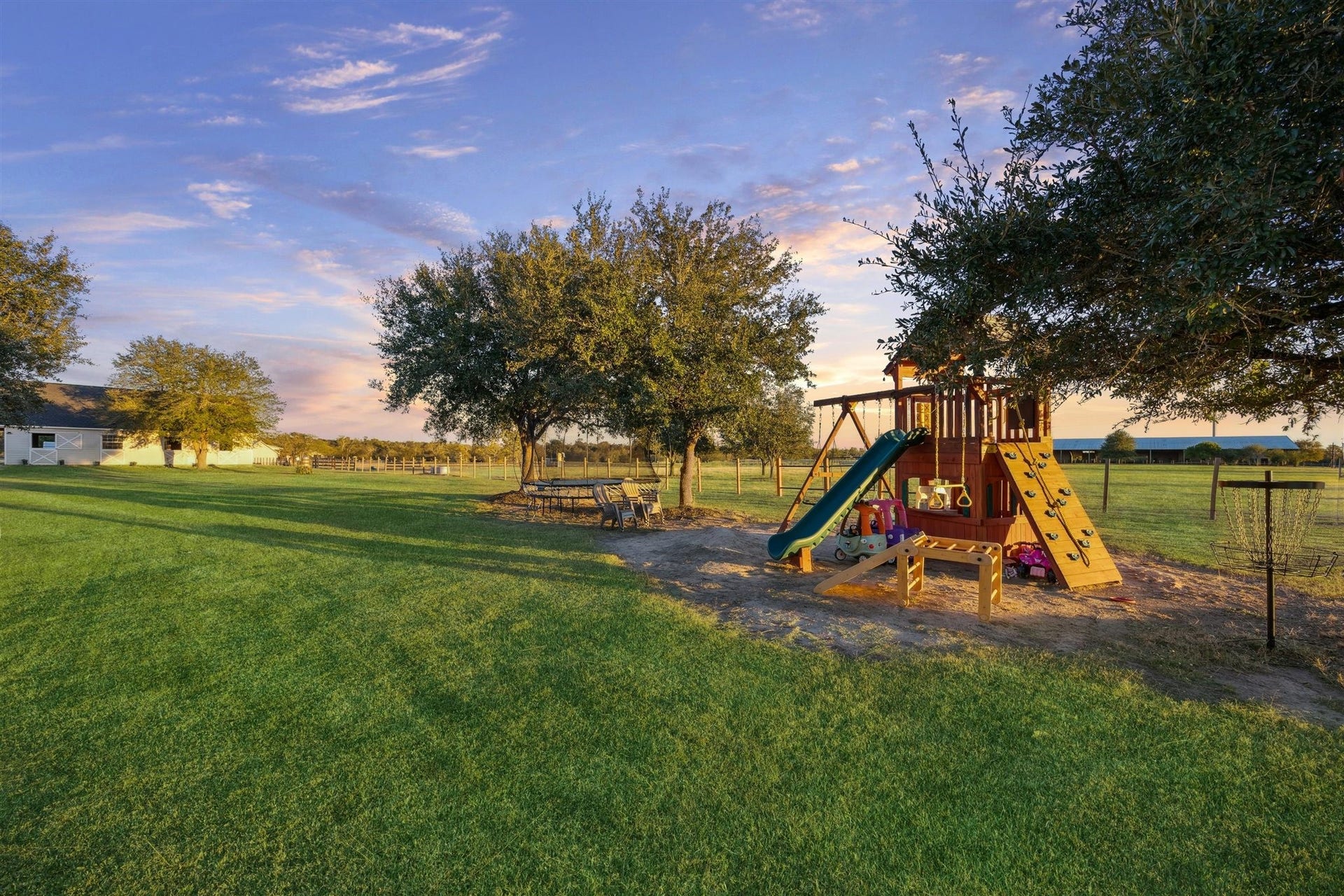 Surrounded by open pasture and shaded by mature trees, this backyard playground is a dream for children. Featuring a wooden playset with rock wall, swings, slide, and monkey bars, plus a picnic table beneath the trees, its built for joyful afternoons and endless imagination, set on land that benefits from a wildlife exemption, blending play with purpose.