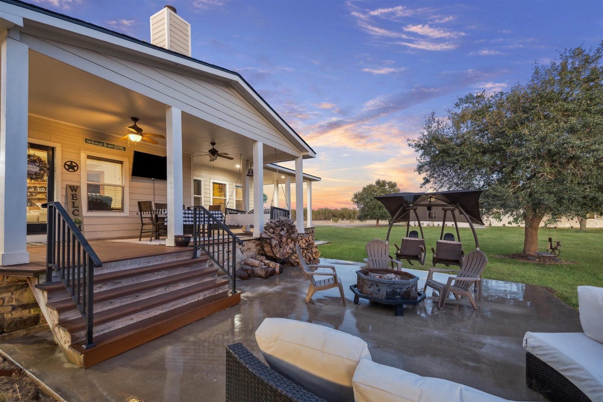 This inviting backyard retreat features a charming firepit lounge surrounded by Adirondack chairs and swings, with a neat stack of firewood ready for the season. The covered porch offers fans, soft lighting, and a cozy dining nook perfect for winding down at sunset or gathering with guests beneath the wide Texas skies.