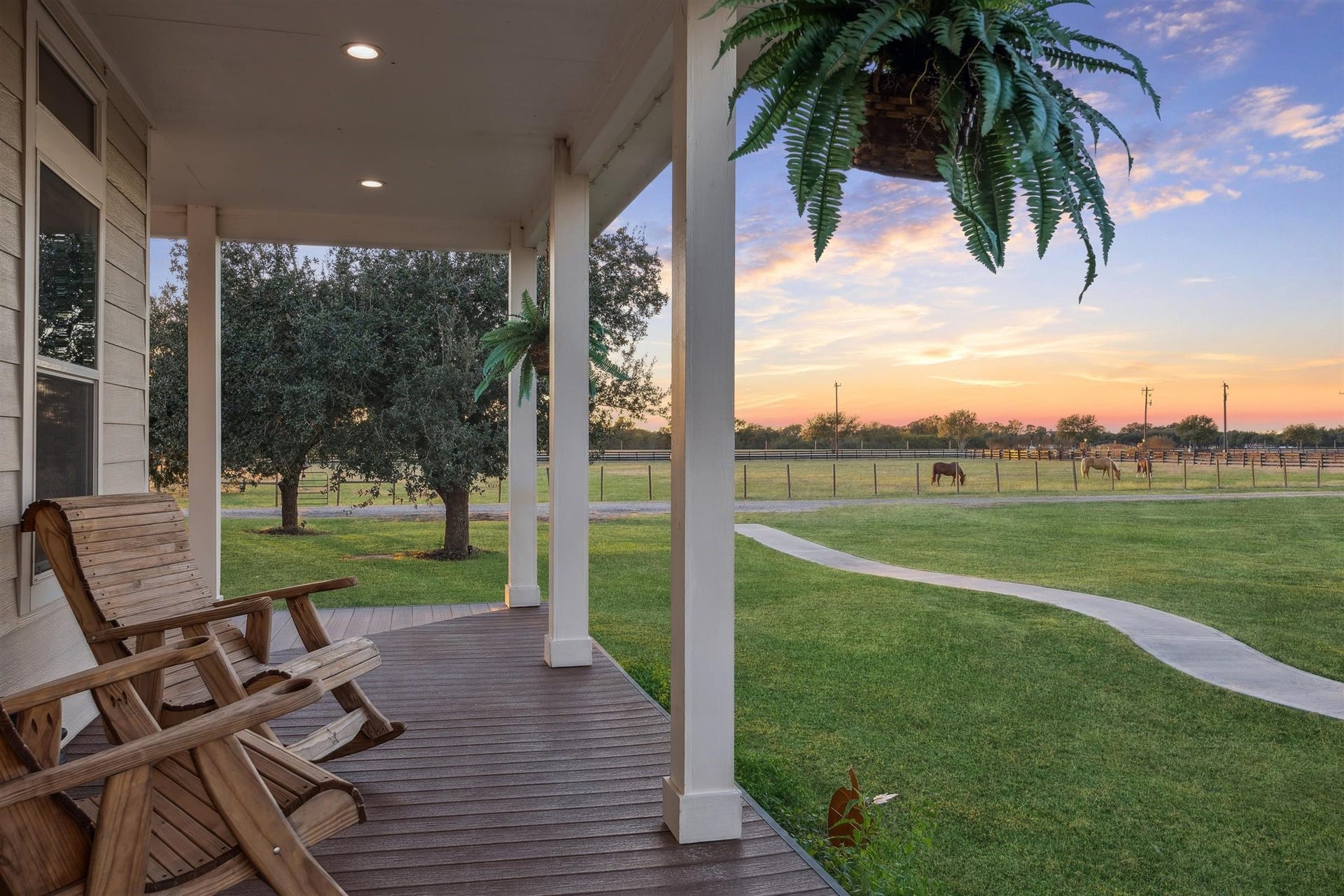 A peaceful evening on the porch offers serene views of grazing horses and sunset-drenched pastures. Wooden rockers invite you to slow down and take in the calm, while the gently curving path winds through lush, fenced acreage dotted with mature trees, land that not only inspires but also qualifies for a wildlife exemption, enhancing both beauty and benefit.