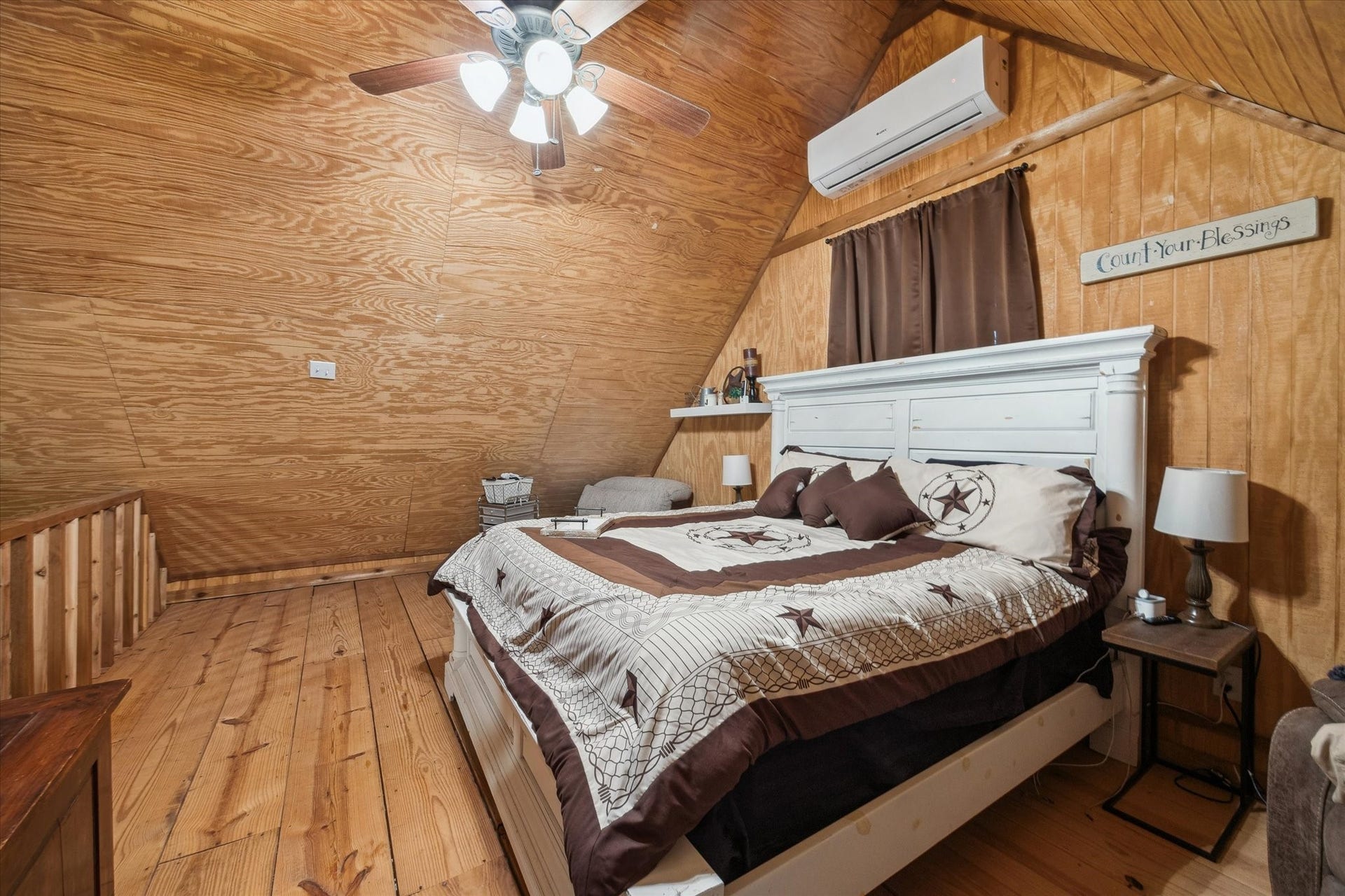 This loft bedroom combines rustic simplicity with modern comfort. Wood walls and vaulted ceilings give it a warm cabin feel, while a ceiling fan and mini-split A/C keep the space cool and inviting all year round.
