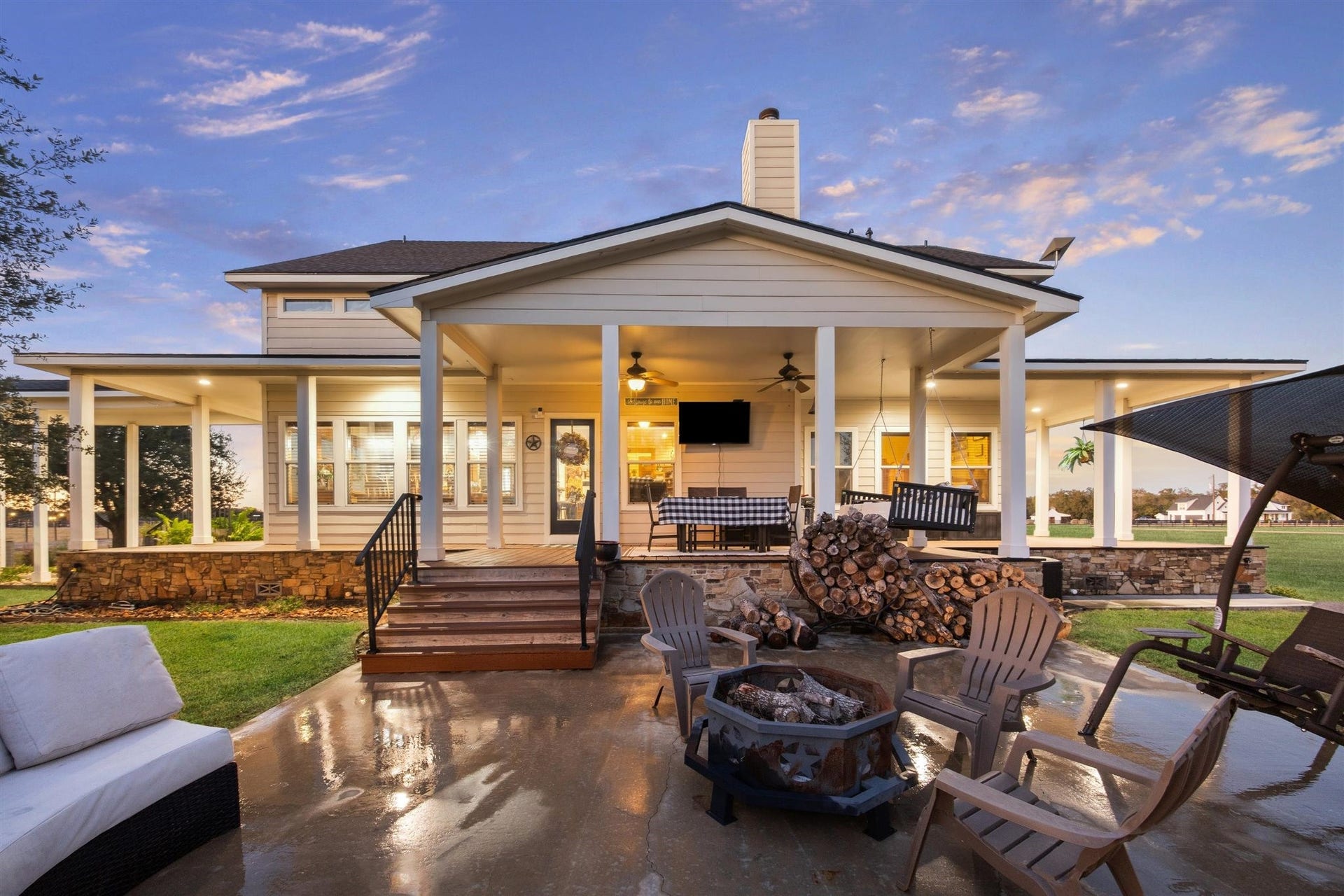 As dusk sets in, the full view of the wraparound porch glows with warmth and welcome. With dual staircases, porch swings, and stacked firewood for the firepit, this backyard is perfectly designed for entertaining or simply relaxing into the calm of a country evening.