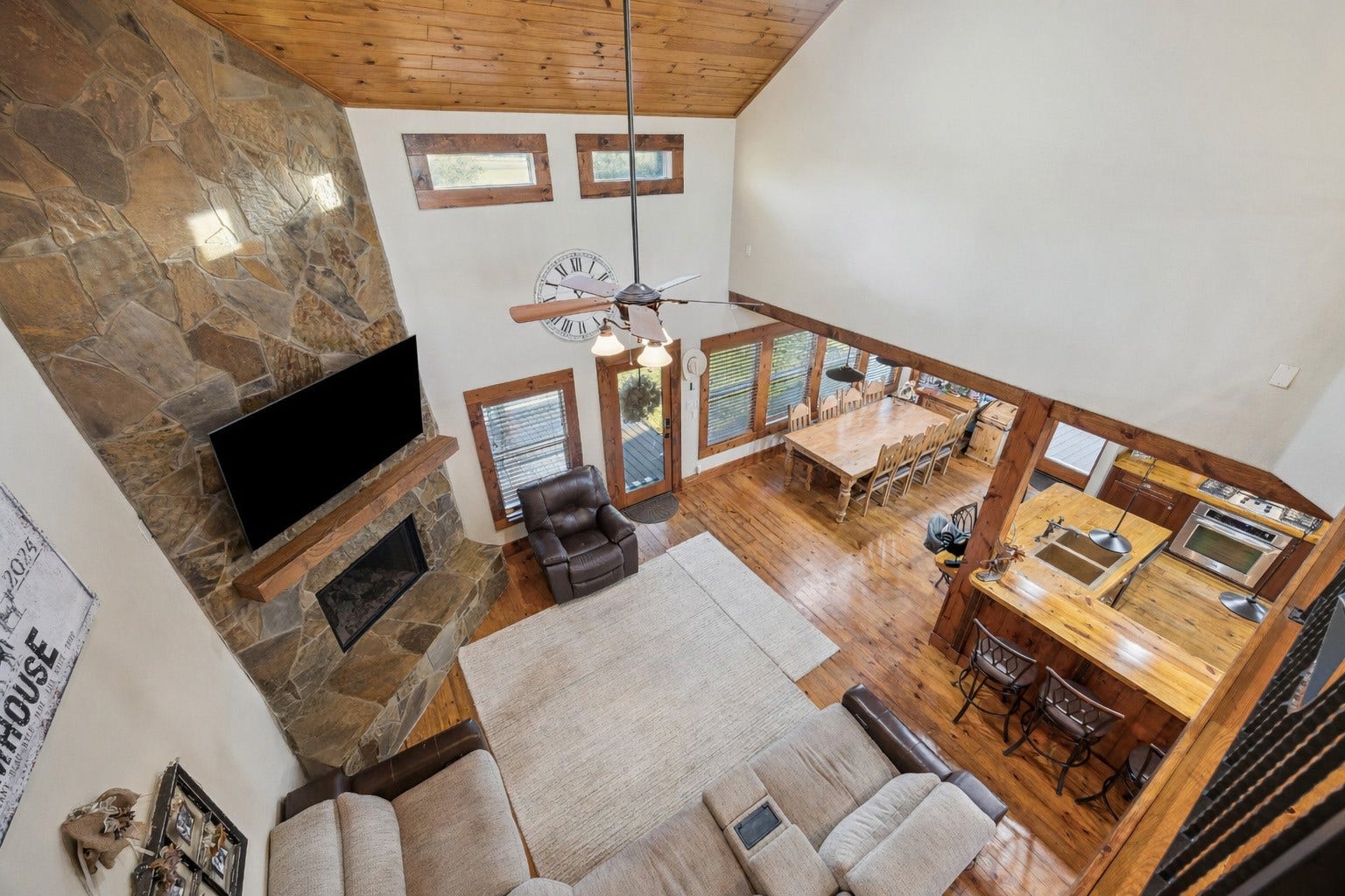 The heart of the home features a grand floor-to-ceiling stone fireplace, cathedral ceilings, warm wood trim, and open views to the dining room and kitchen. A true gathering space with cozy touches and a seamless connection to indoor-outdoor living.