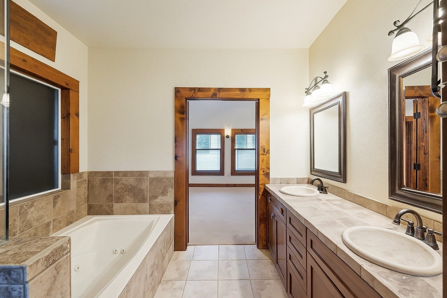 Unwind in this spa-style bathroom featuring a jetted tub, dual vanities with stone counters, and warm wood accents. The open doorway leads directly back into the relaxing primary bedroom, seamlessly connecting comfort and luxury.