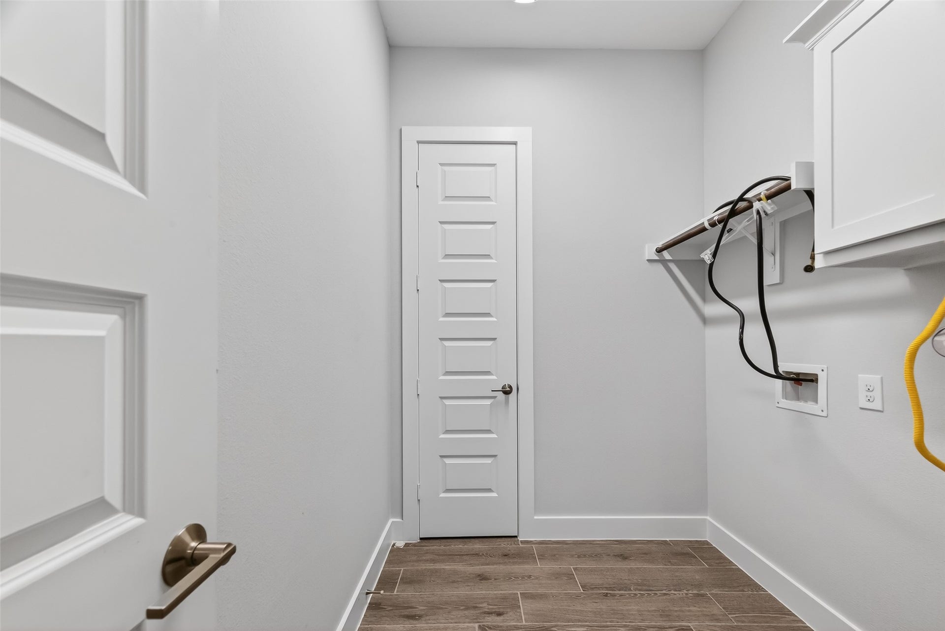 3415 Mckinney Ridge Drive, Houston, TX 77059 This photo shows a utility room with light gray walls and wood-look tile flooring. It features a white door, cabinets, and connections for a washer and dryer, with an overhead shelf for storage.